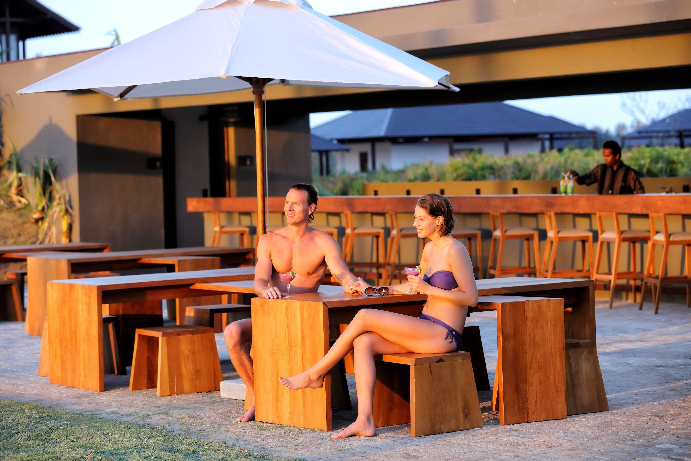 Un couple est assis à une table en bois sous un parasol blanc. On aperçoit en arrière-plan un bar et des tables à l'extérieur.