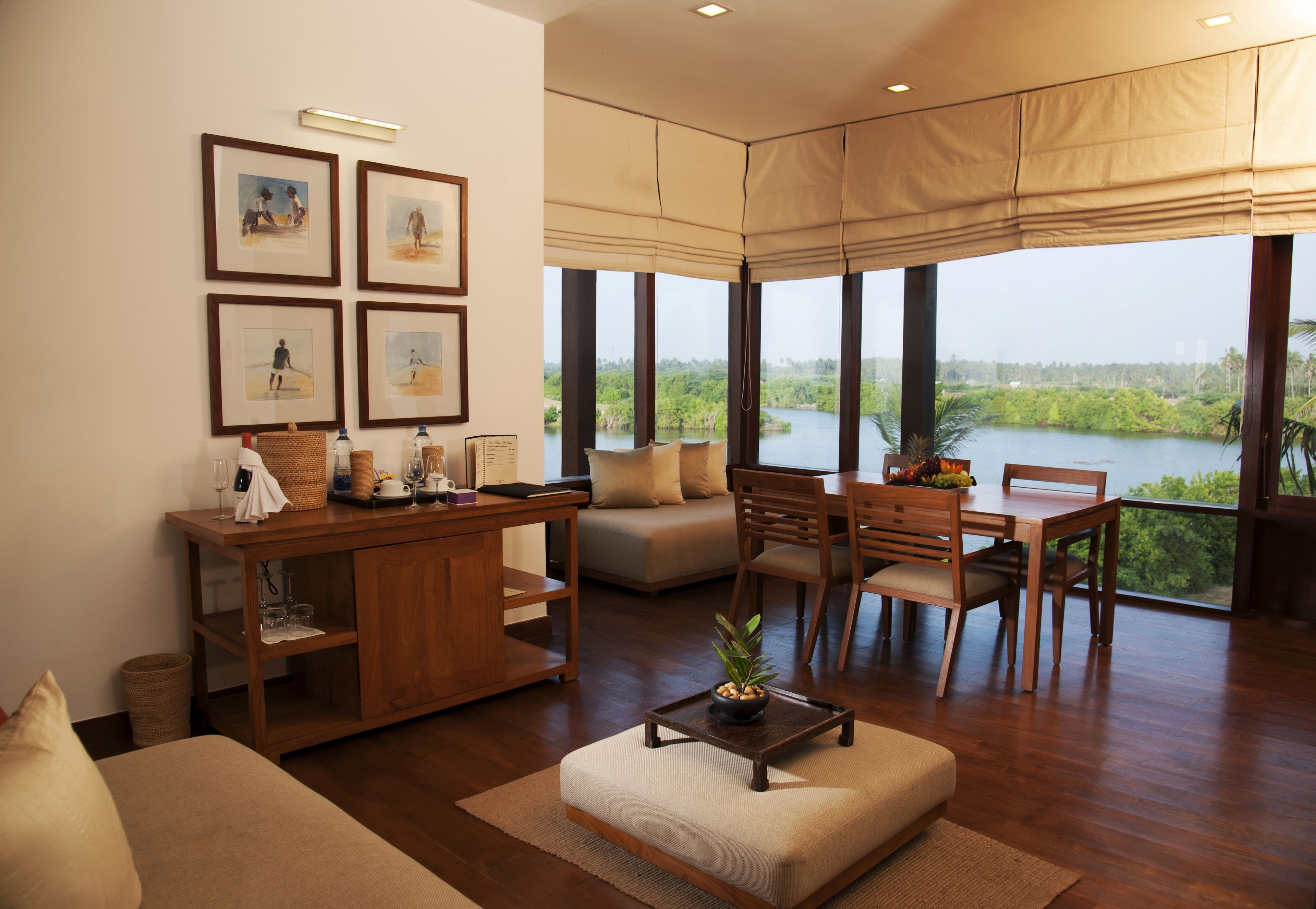 A modern living room with a wooden coffee table, a sofa, and a dining area with chairs. There are framed pictures on the wall and a large window with a view of a body of water.