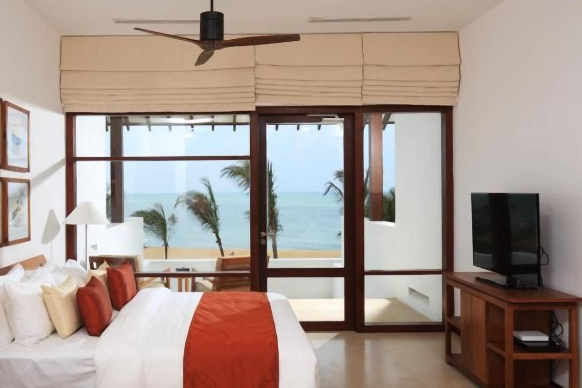Habitación de hotel con cama, televisor, ventilador de techo y ventana grande con vista a la playa.