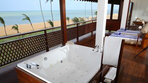Une terrasse en bois avec une grande baignoire, un coin salon avec des coussins à rayures et une vue sur la plage et l’océan.
