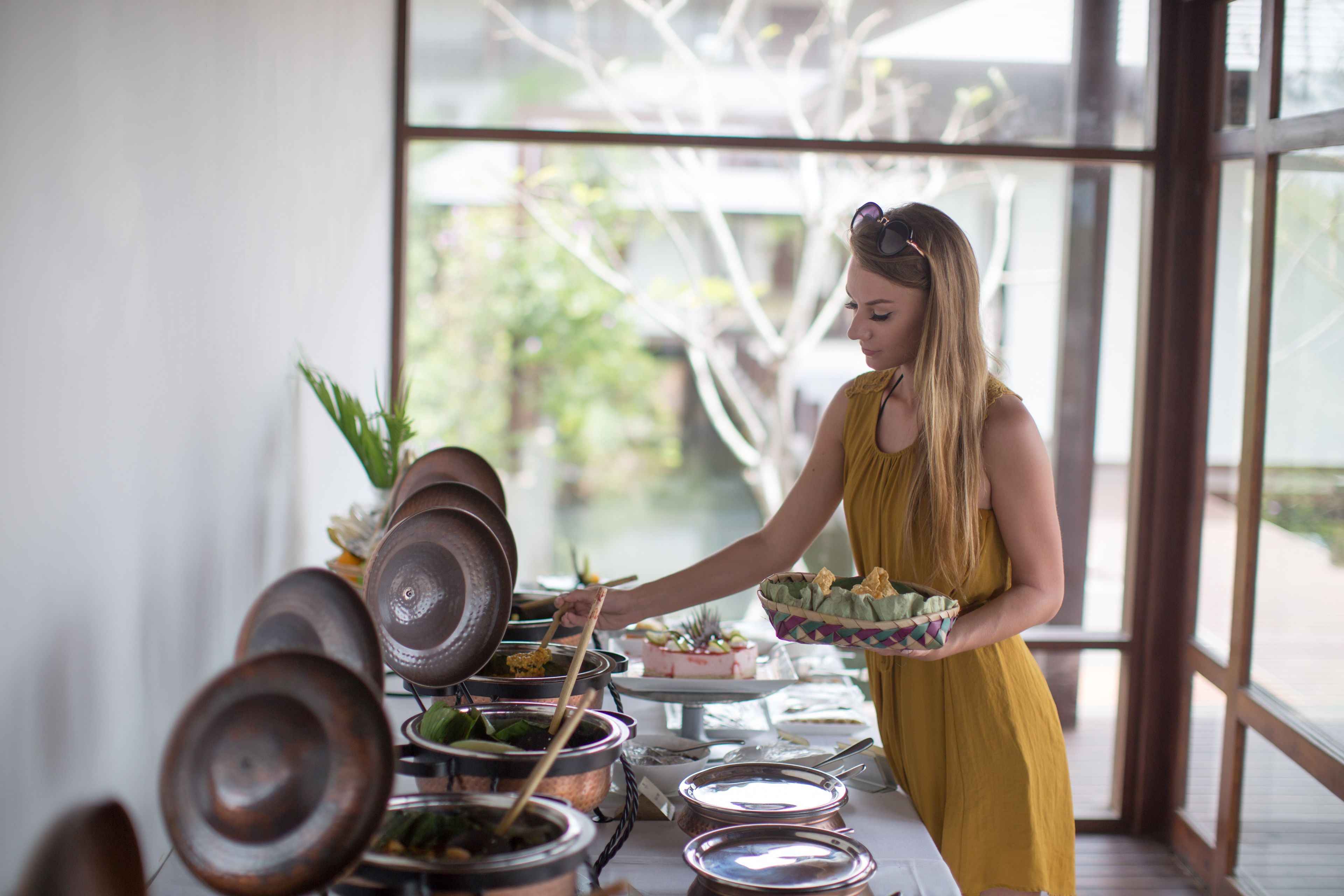 Eine Frau im gelben Kleid serviert Speisen an einem Buffet mit verschiedenen Gerichten und Besteck.