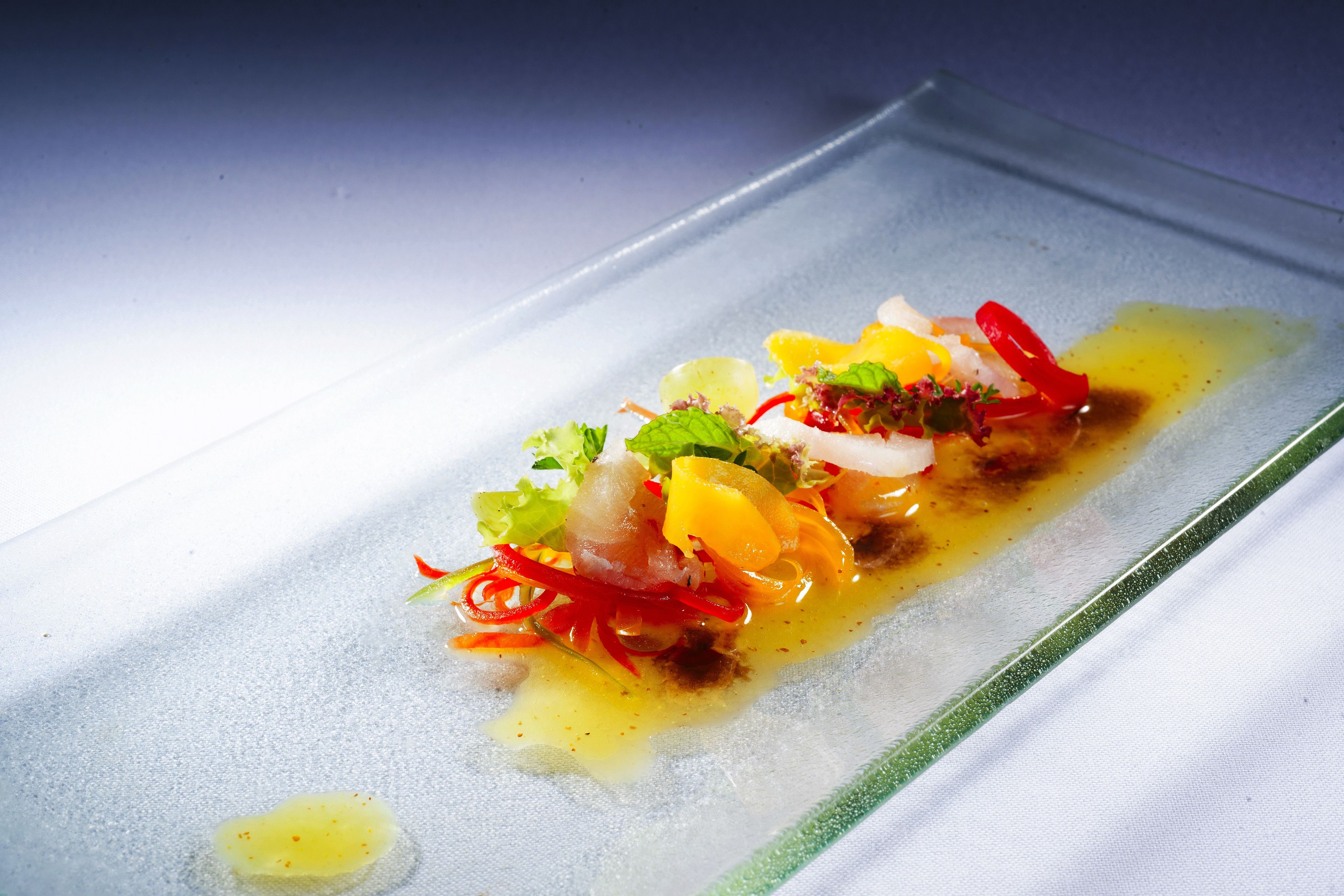 A glass plate with a colorful dish, possibly a salad or appetizer, with various vegetables and a drizzle of sauce.