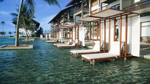 Un complexe hôtelier doté de chaises longues confortables au bord de la piscine, d’une promenade bordée de palmiers et d’un bâtiment avec des balcons offrant une vue sur l’eau.