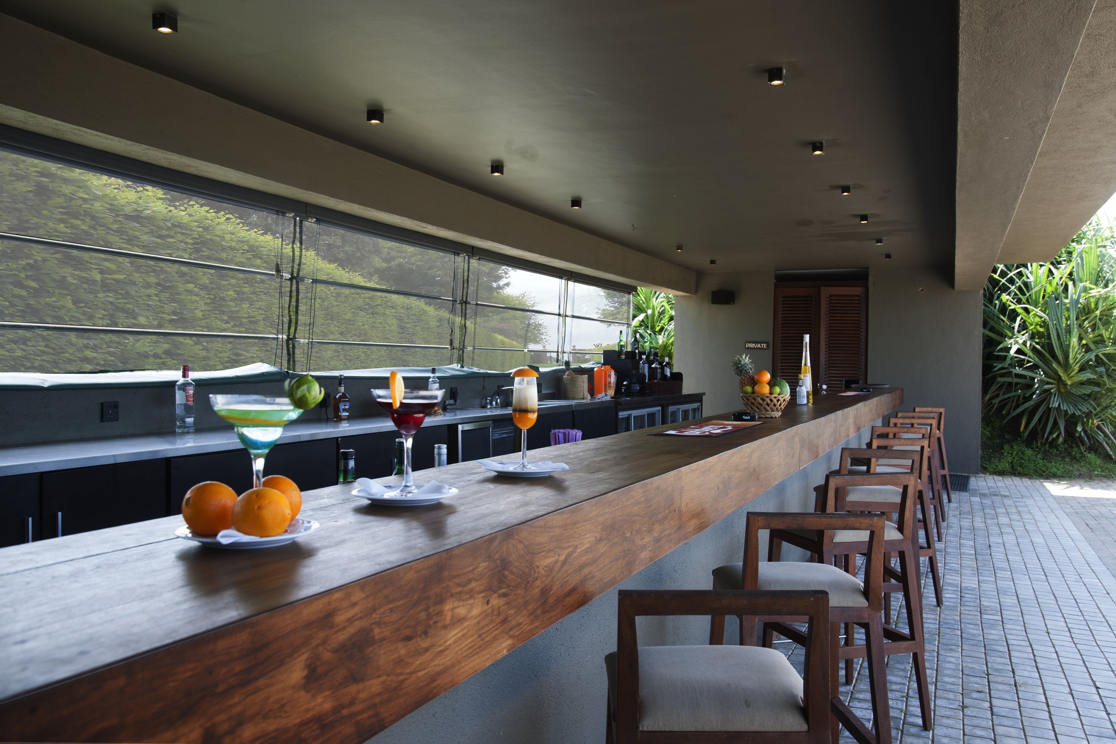 A modern bar area with a long wooden counter, bar stools, and a view of greenery through large windows.