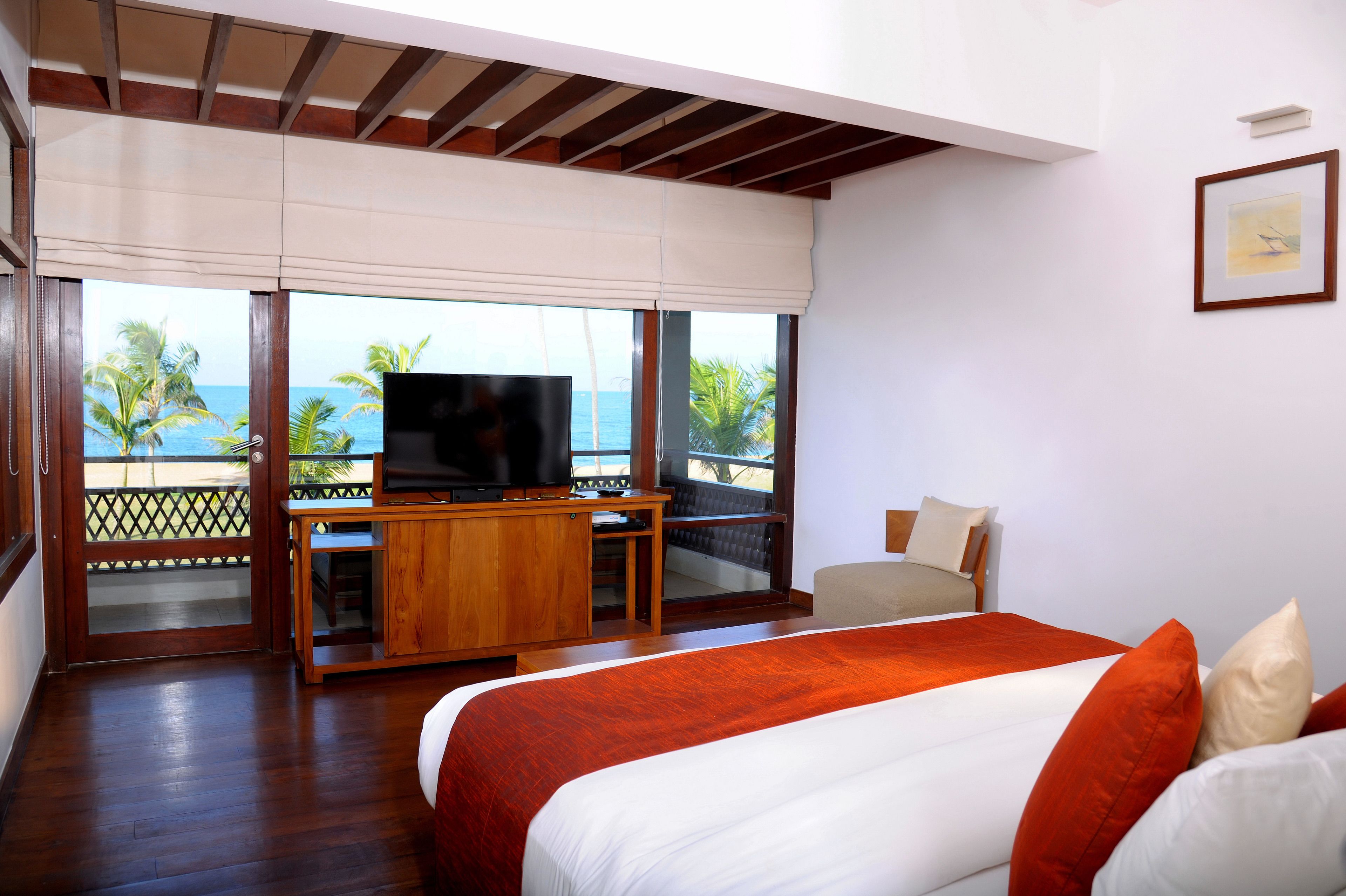 A hotel room with a large bed, a TV on a wooden cabinet, and a balcony with a view of the ocean.