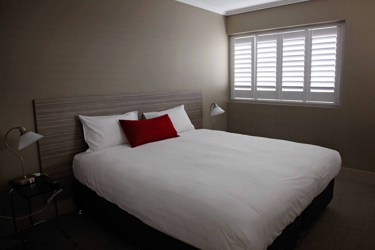A neatly made bed with white and red pillows, a bedside lamp, and a window with blinds.