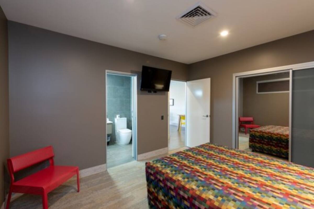 A bedroom with a bed, a red bench, a television, and a bathroom visible through an open door.