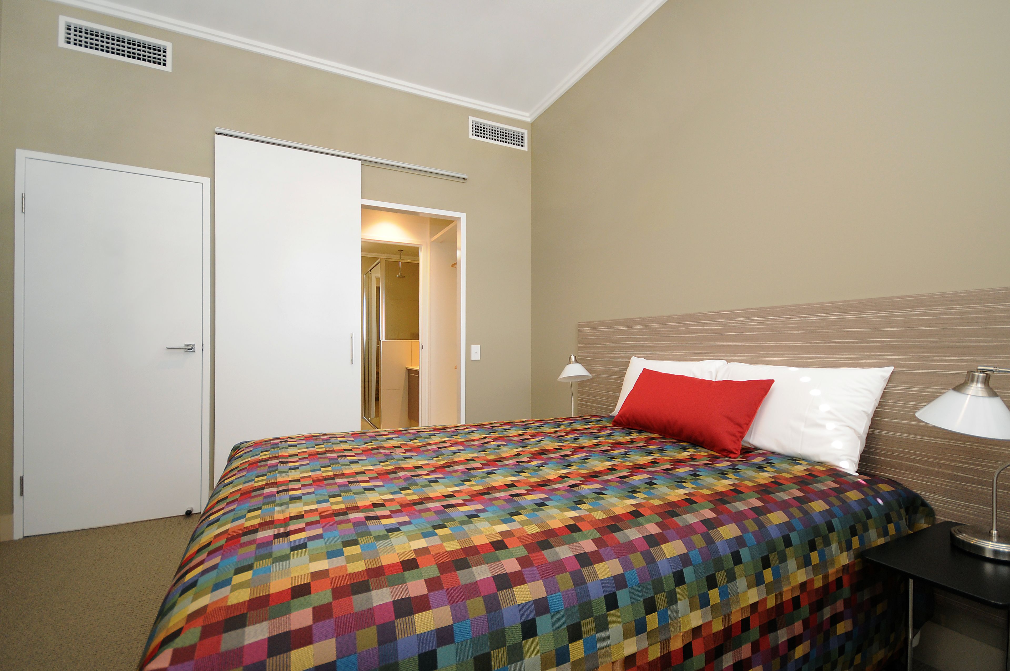 Habitación de hotel con una cama grande, una mesita de noche con lámpara, una puerta y un pasillo que lleva a otra habitación.