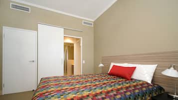 Habitación de hotel con una cama grande, una mesita de noche con lámpara, una puerta y un pasillo que lleva a otra habitación.