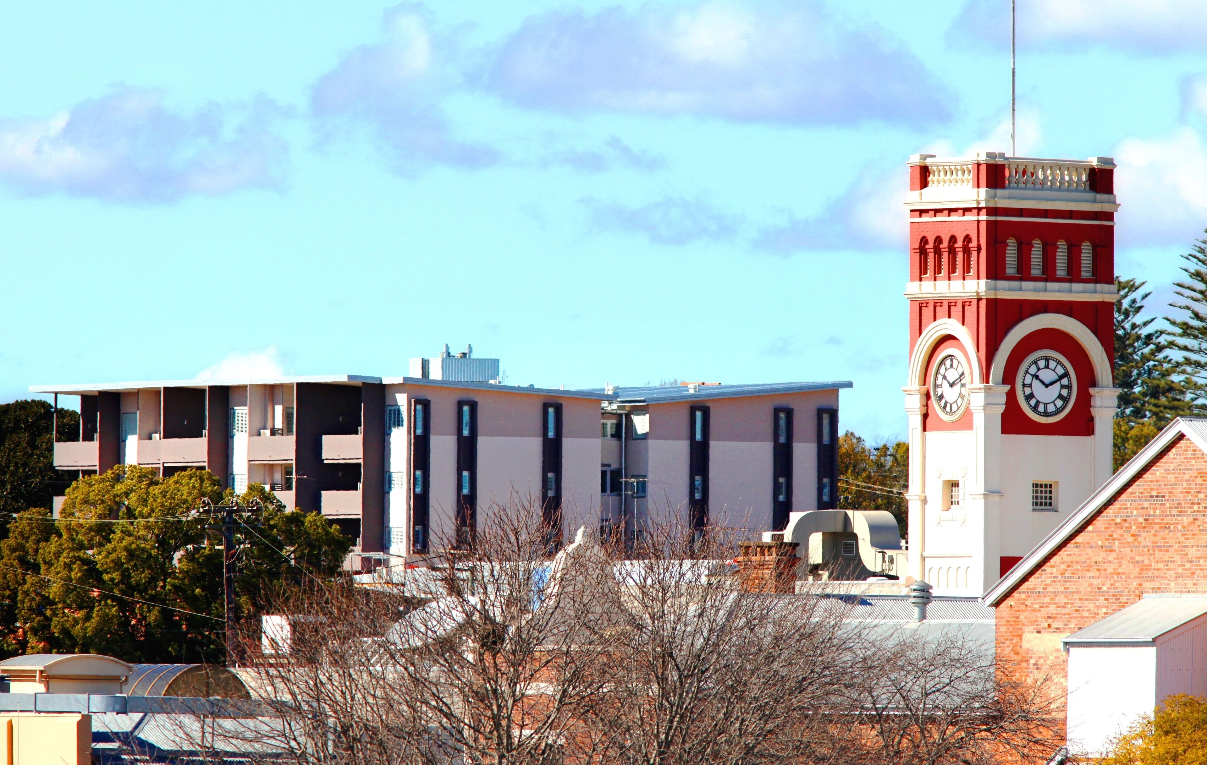 A red and white clock tower with a clock face, surrounded by modern buildings with balconies and a clear blue sky.
