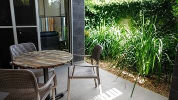 An outdoor patio with a round table and two chairs, surrounded by lush greenery and a brick wall.