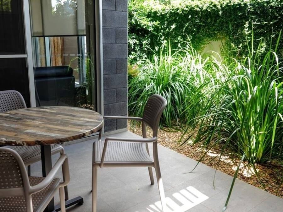 An outdoor patio with a round table and two chairs, surrounded by lush greenery and a brick wall.