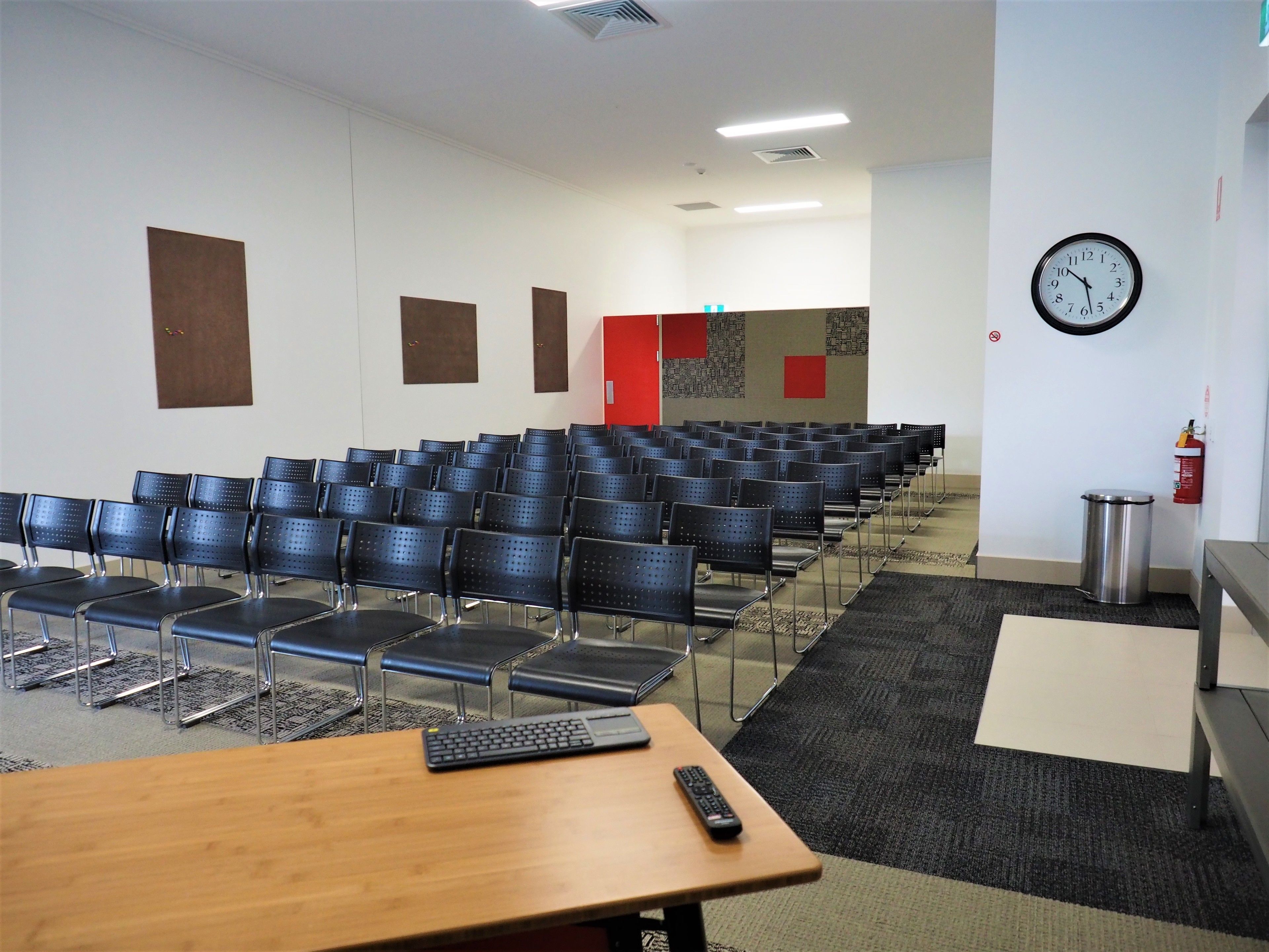 A conference room with rows of chairs, a wooden table with a keyboard and remote, a clock, and a fire extinguisher.