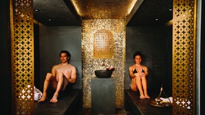 A spa room with two people seated on benches, a decorative wall, and a small table with a bottle and a bowl. - Hepburn Spa Retreat (Hepburn Springs)