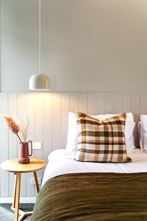 A neatly made bed with a plaid pillow, a small wooden side table with a vase, and a wall-mounted lamp. - Hepburn Spa Retreat (Hepburn Springs)
