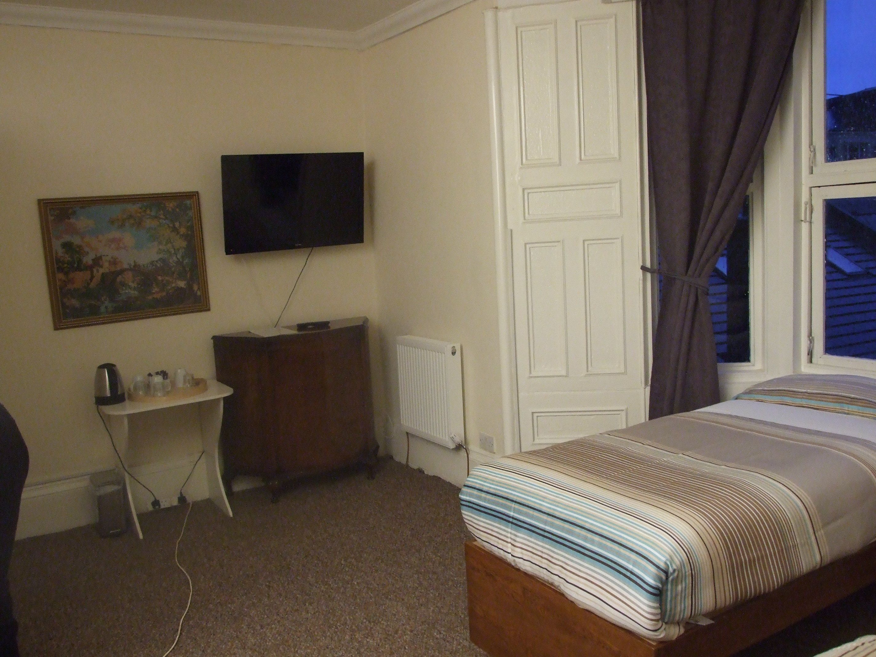 A bedroom with a bed, a TV mounted on the wall, a desk, and a radiator.