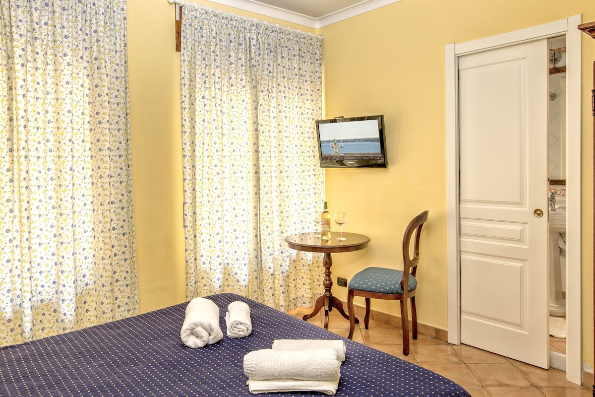 Habitación de hotel con cama, una silla, una mesita con una botella y un vaso, y un televisor en la pared.