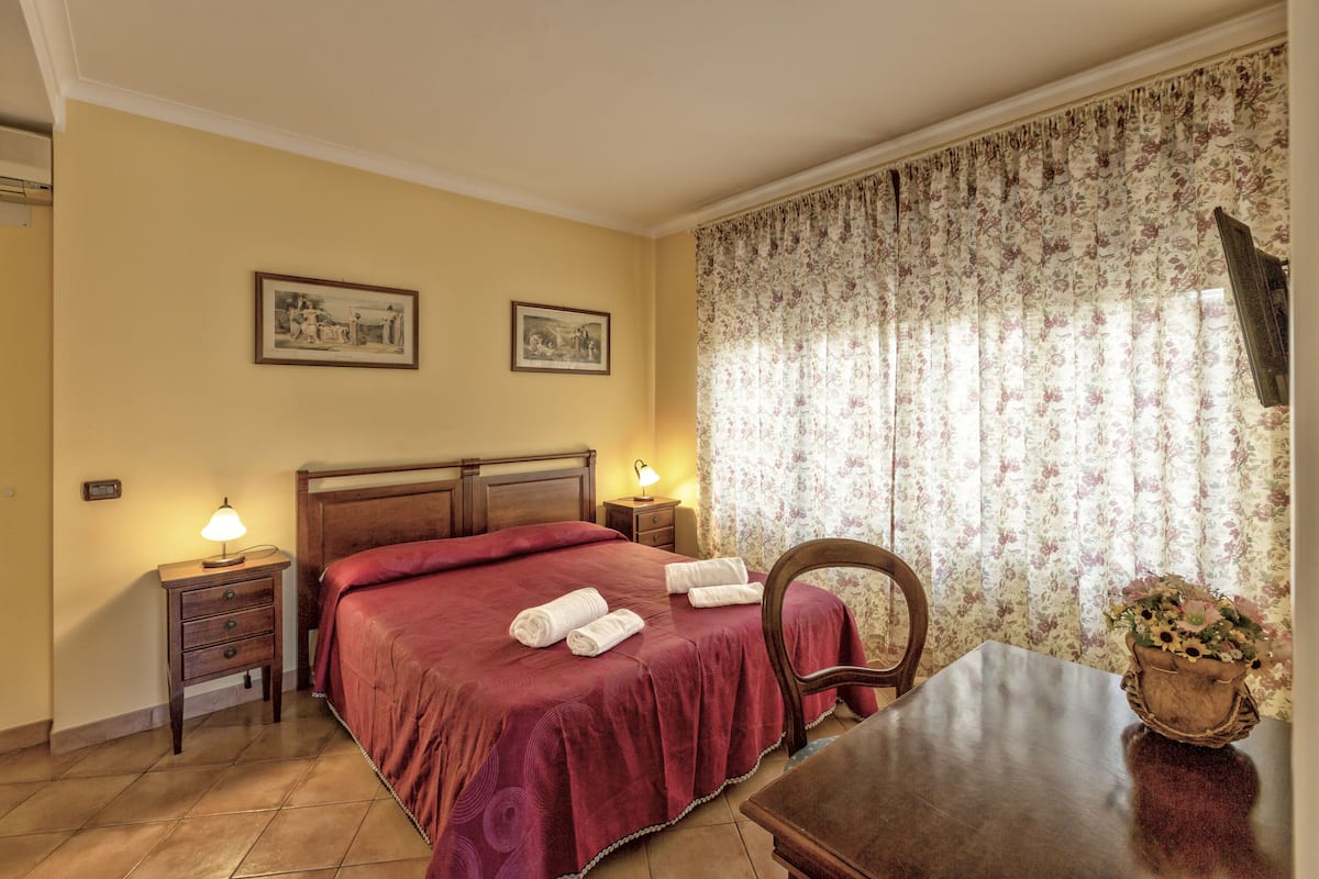 Habitación de hotel con una cama, una mesita de noche con una lámpara, una mesa con un jarrón de flores y dos cuadros en la pared.