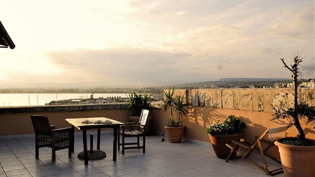 A terrace with a table and chairs, potted plants, and a view of a body of water and distant landscape. - Bed & Breakfast Armonia (Syracuse)