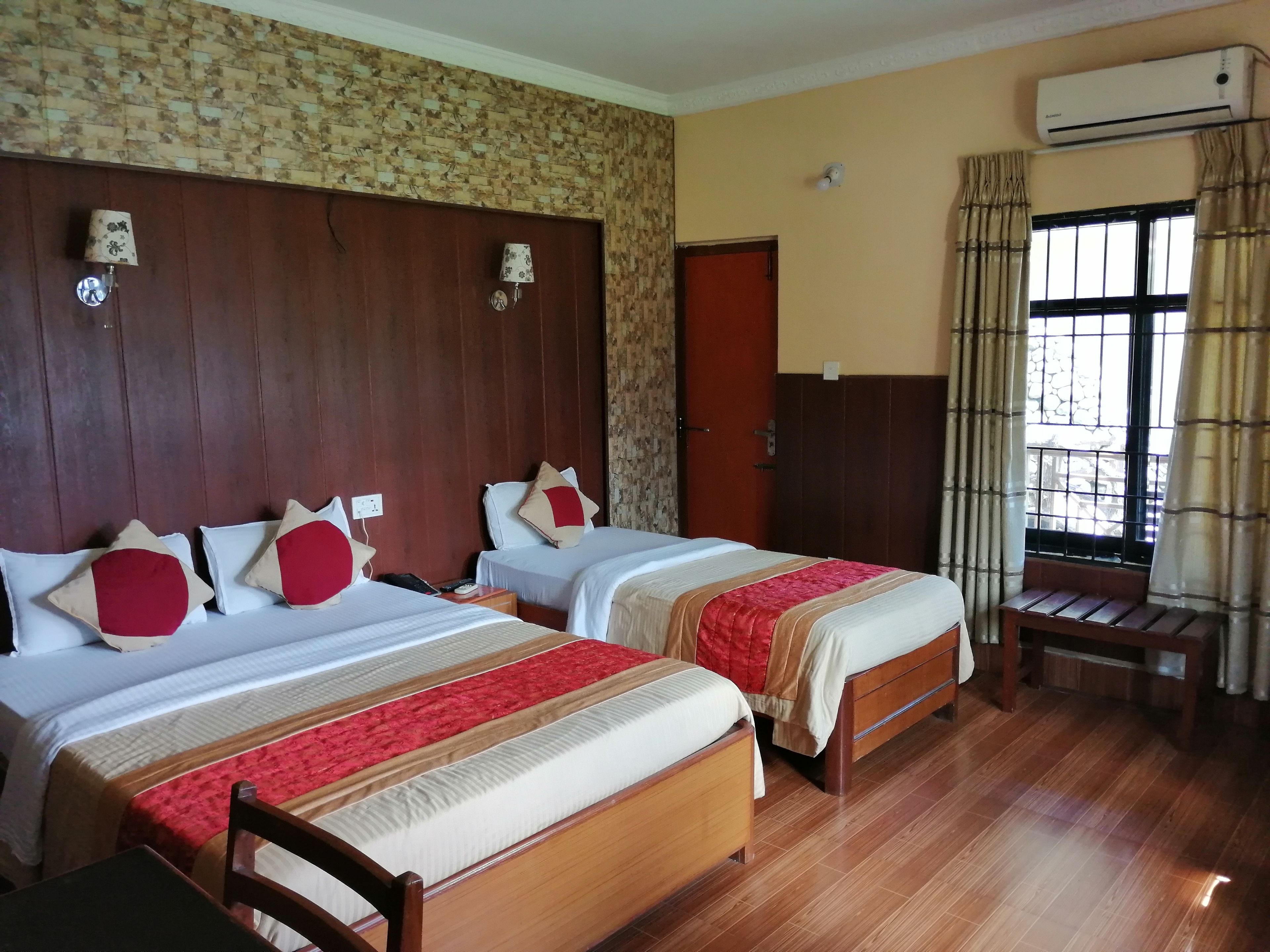 A hotel room with two beds, wooden headboard, a wooden bench, an air conditioning unit, and a window with curtains.