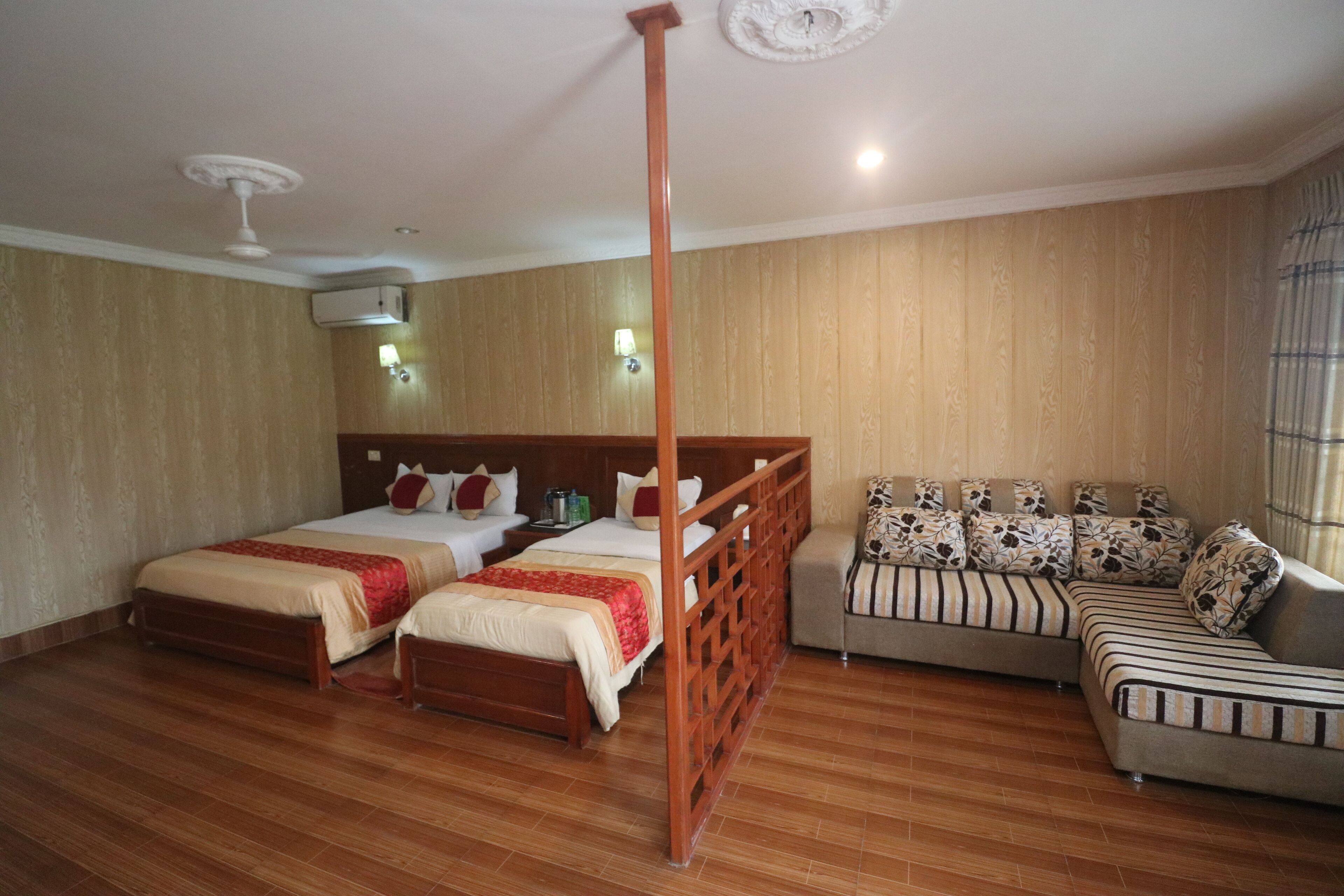 A hotel room with two single beds, a sofa, wooden flooring, a ceiling fan, and an air conditioning unit.