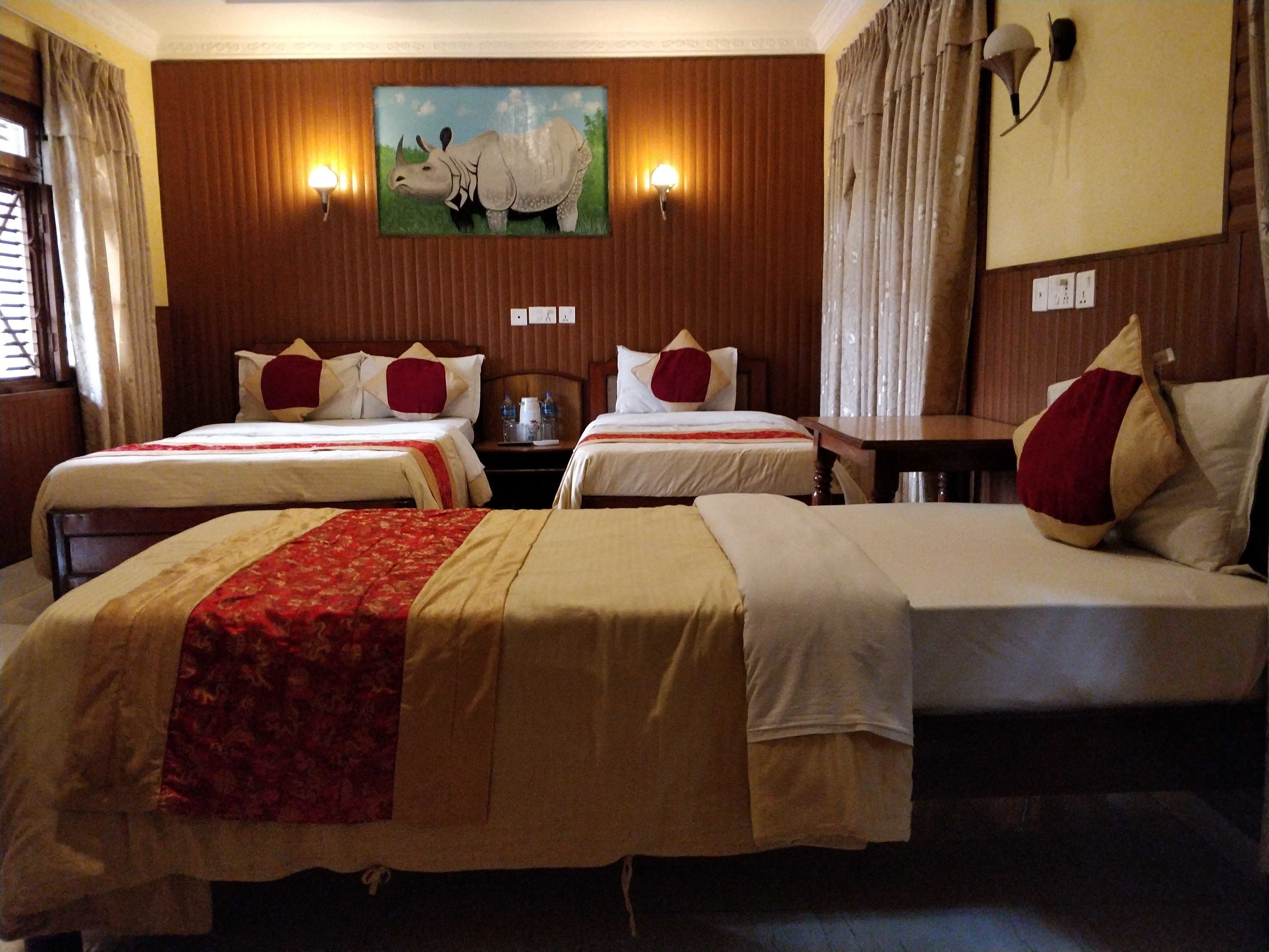 A hotel room with two single beds and one double bed, wooden paneling on the walls, a painting of rhinoceroses, and a nightstand with a lamp.