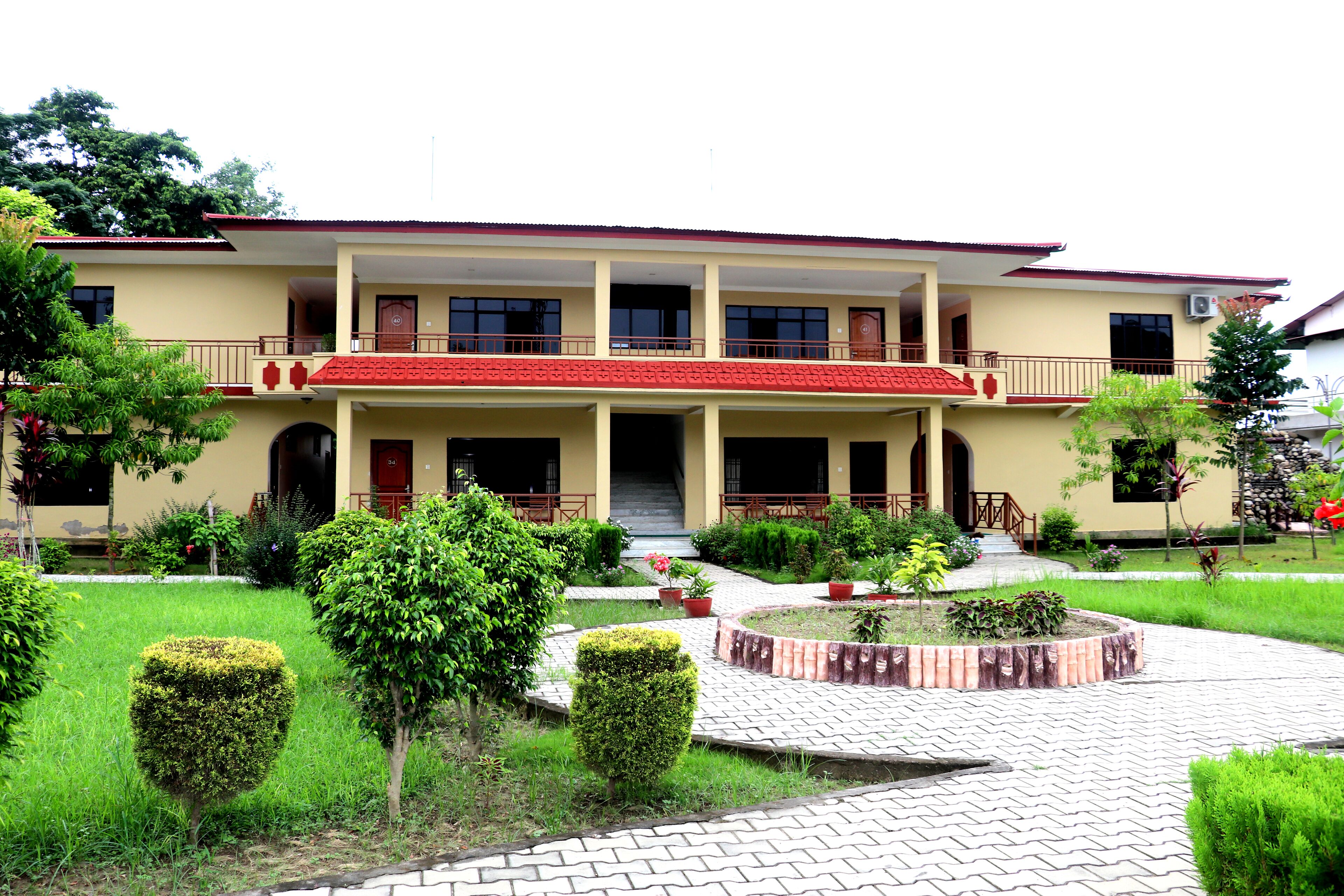 Un edificio de dos pisos con techo rojo y paredes color beige, con una entrada central que incluye una escalera, rodeado de jardines cuidados y un sendero pavimentado.