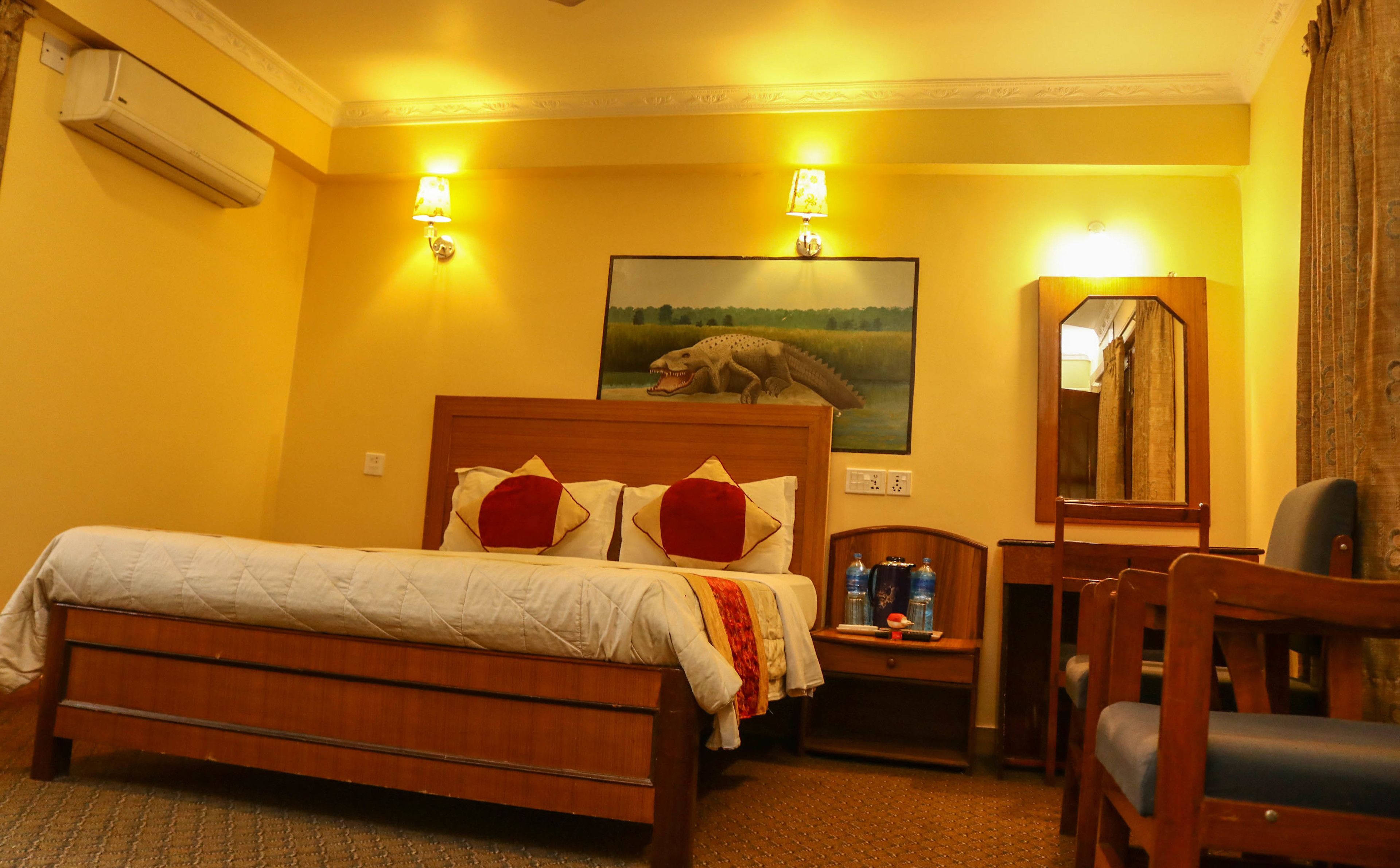A hotel room with a wooden bed, two wall-mounted lamps, a painting of a dinosaur, a mirror, and a small table with a bottle and a can.