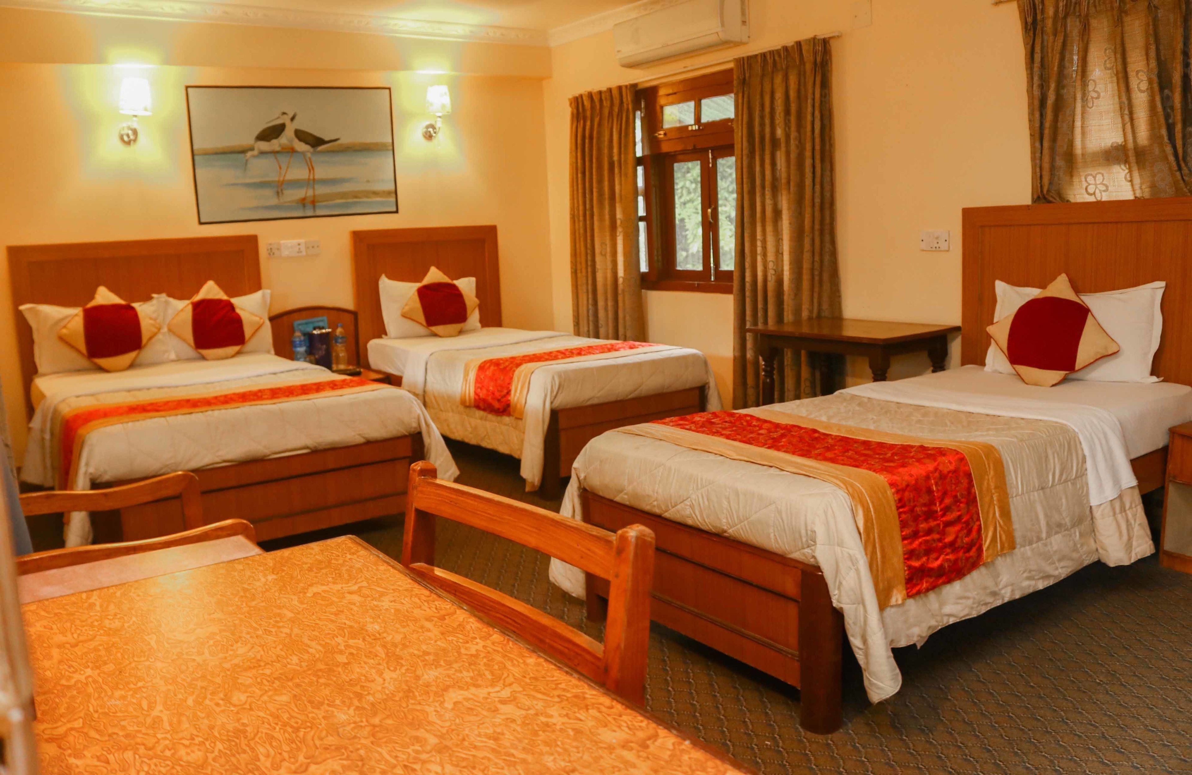 A hotel room with three single beds, each with a wooden headboard and white bedding. There is a wooden table in the foreground, and a window with curtains on the right side.