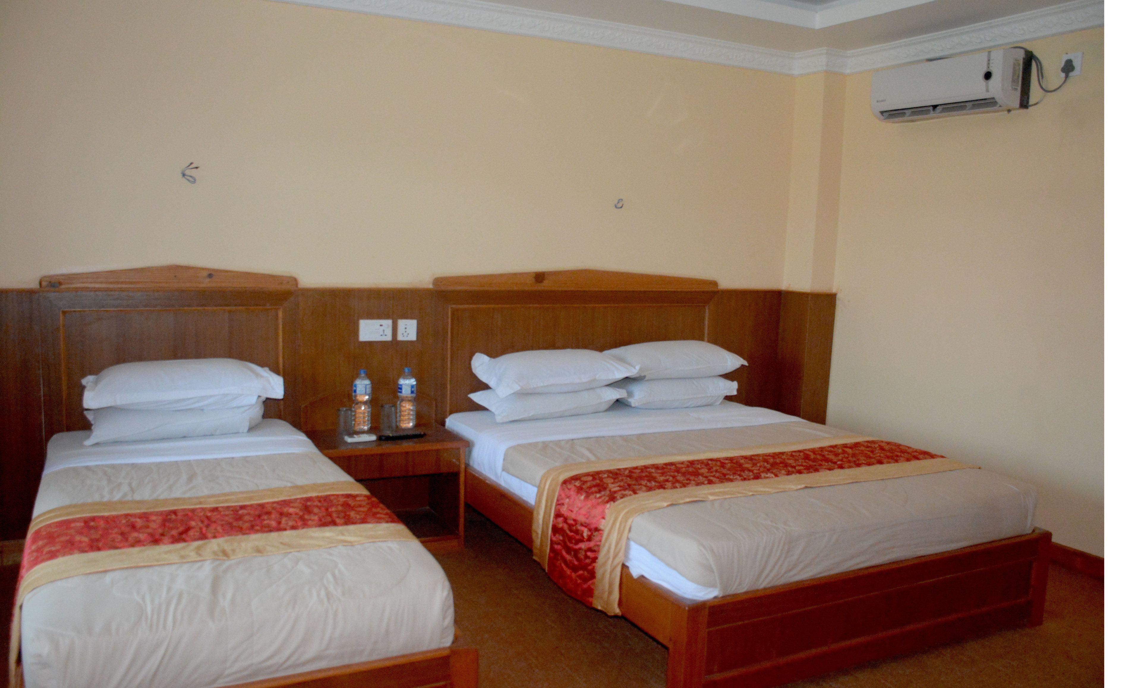 Une chambre d’hôtel avec deux lits simples, des têtes de lit en bois, du linge de lit blanc, un couvre-lit rouge et beige, une table de chevet avec deux bouteilles d’eau, une télécommande et un appareil de climatisation fixé au mur.