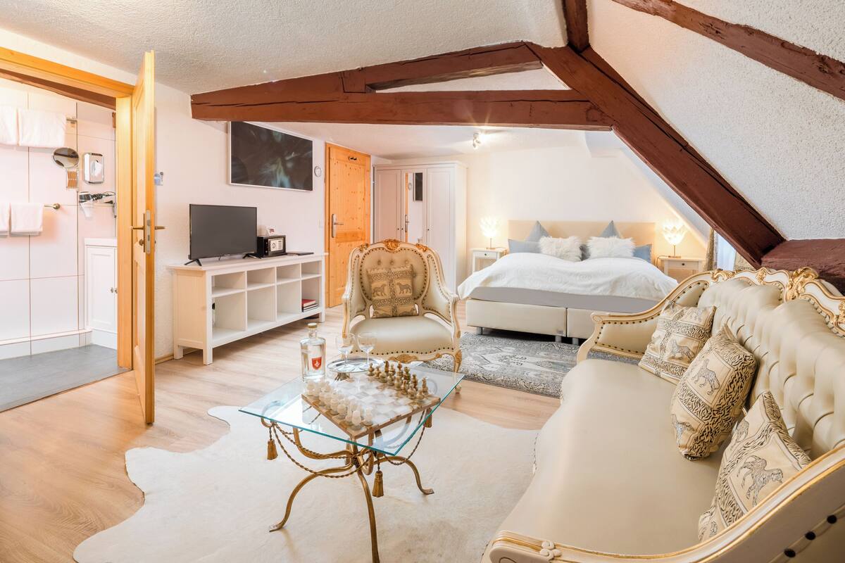 Een ruime kamer met een glazen salontafel, een comfortabele bank, een klassieke fauteuil en een keurig opgemaakt bed.