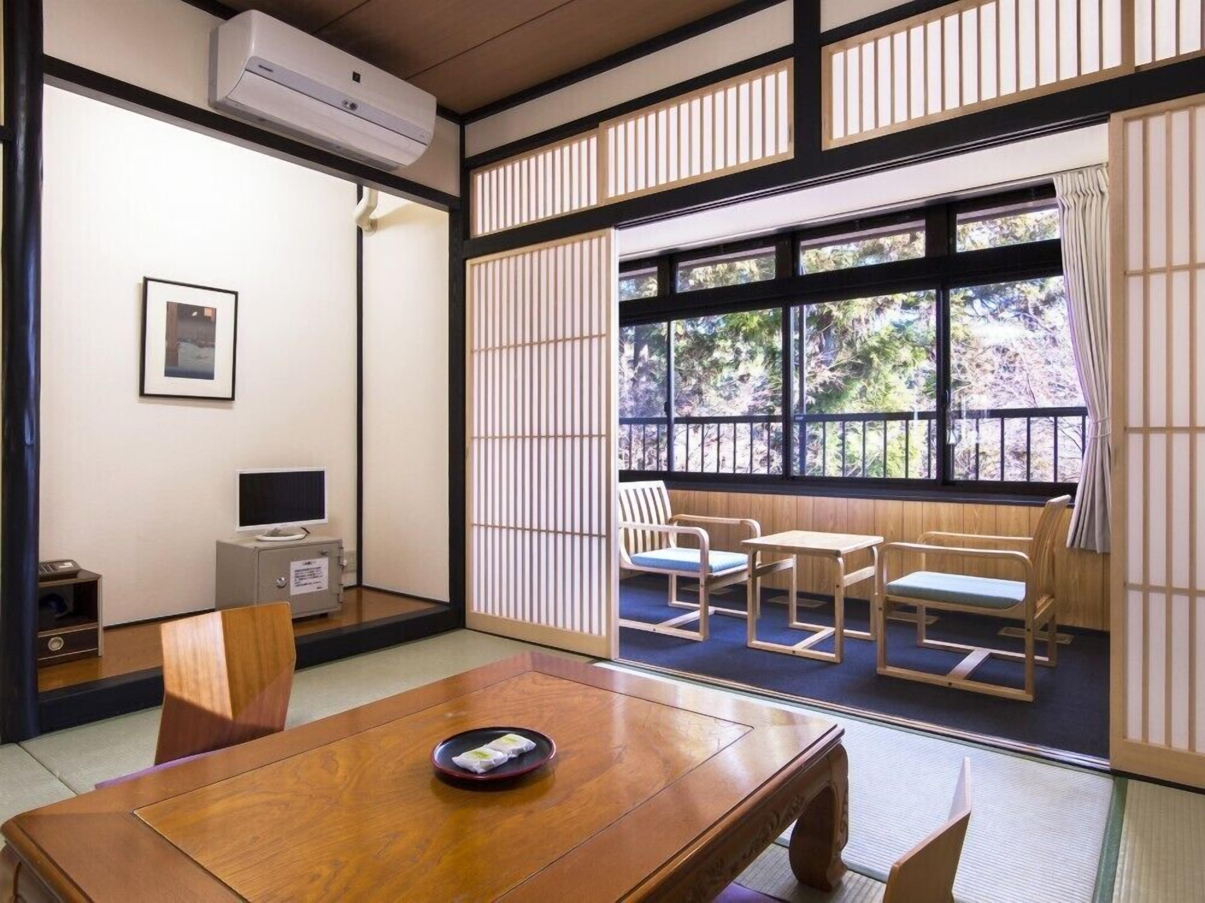 A traditional Japanese room with tatami flooring, a low table with a cushion, a television, and a sliding door leading to a balcony with a view of trees.