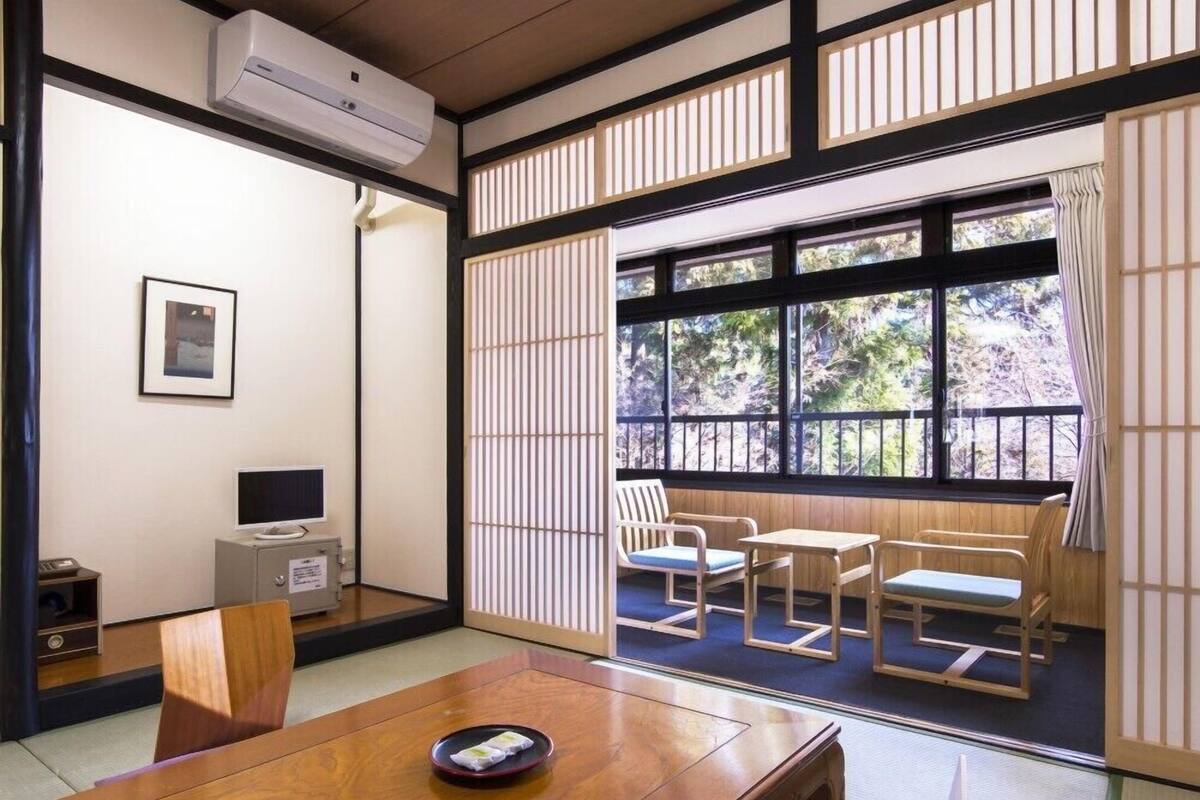 Et traditionelt japansk rum med tatamimåtter, et lavt bord med pude, et fjernsyn og en skydedør, der fører ud til en balkon med udsigt over træer.