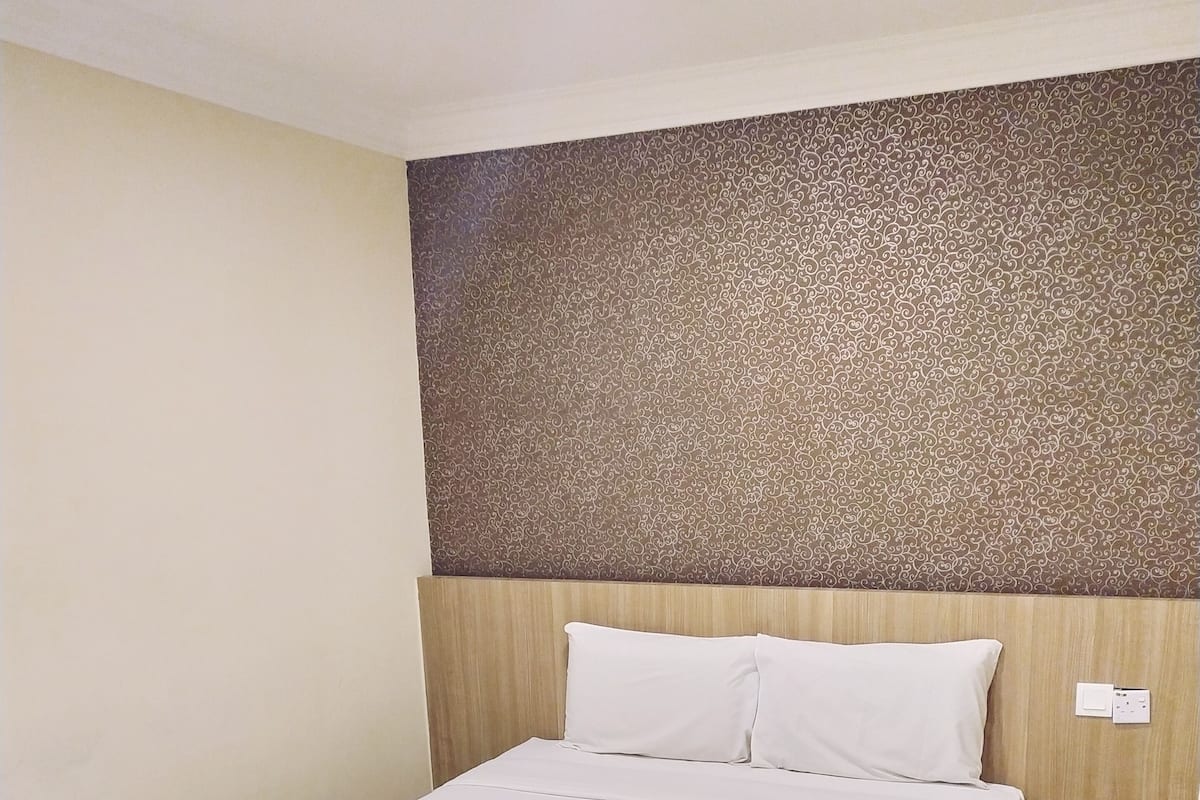 Una cama bien hecha con sábanas blancas y una toalla enrollada, cabecera de madera y pared con estampado.