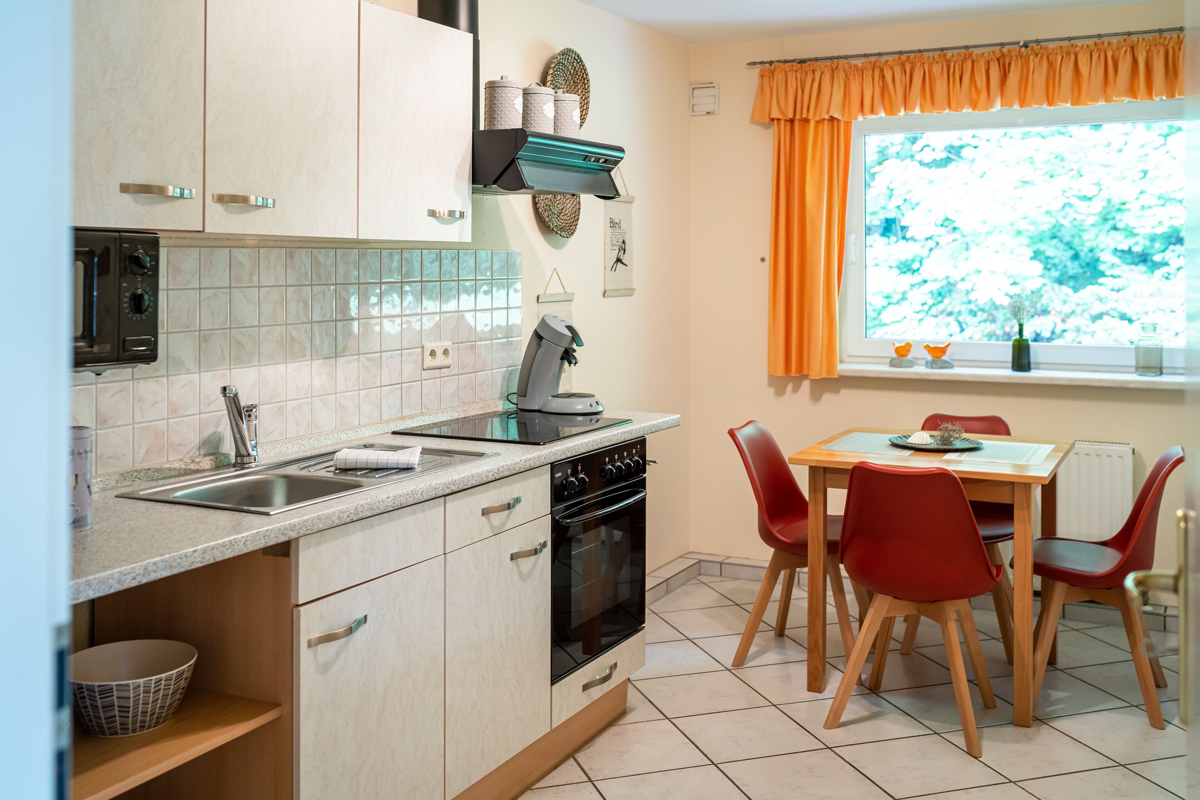 Una cocina con estufa, pileta y alacenas. Se observa una mesa de comedor con sillas y un ventanal con cortinas.