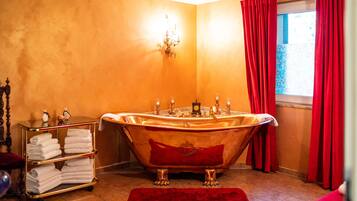 Habitación con una bañera dorada, una alfombra roja, un estante con toallas y una ventana con cortinas rojas.