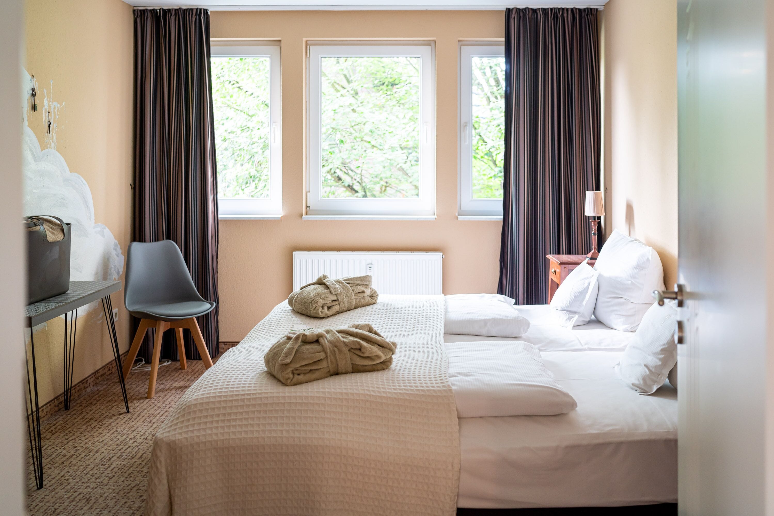 A hotel room with a bed, a chair, a table, and three windows.