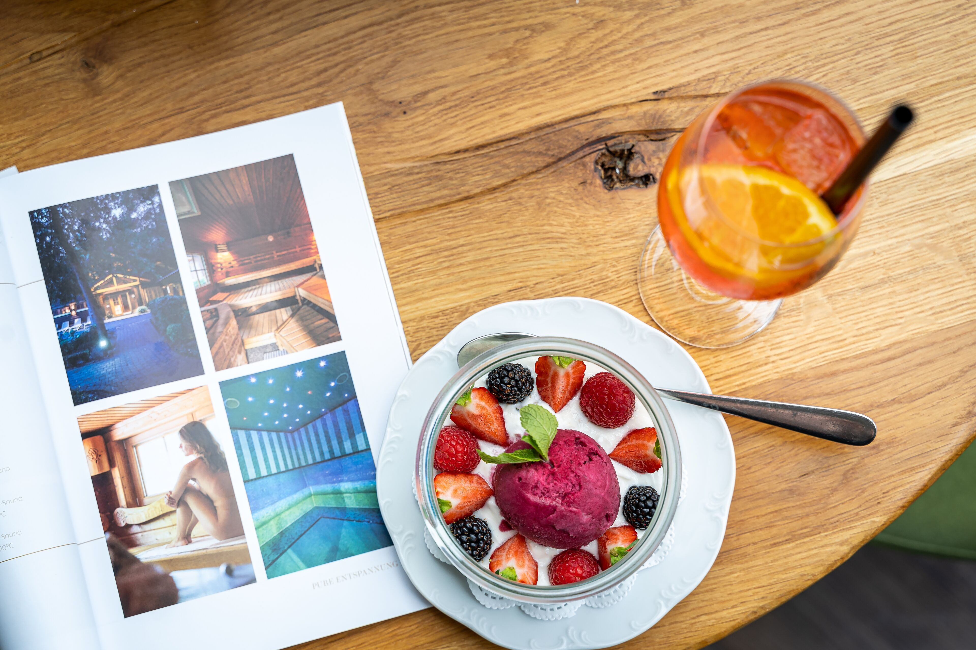 Sobremesa com morangos e amoras, um copo de chá gelado com uma fatia de laranja e uma revista com fotografias sobre uma mesa de madeira.