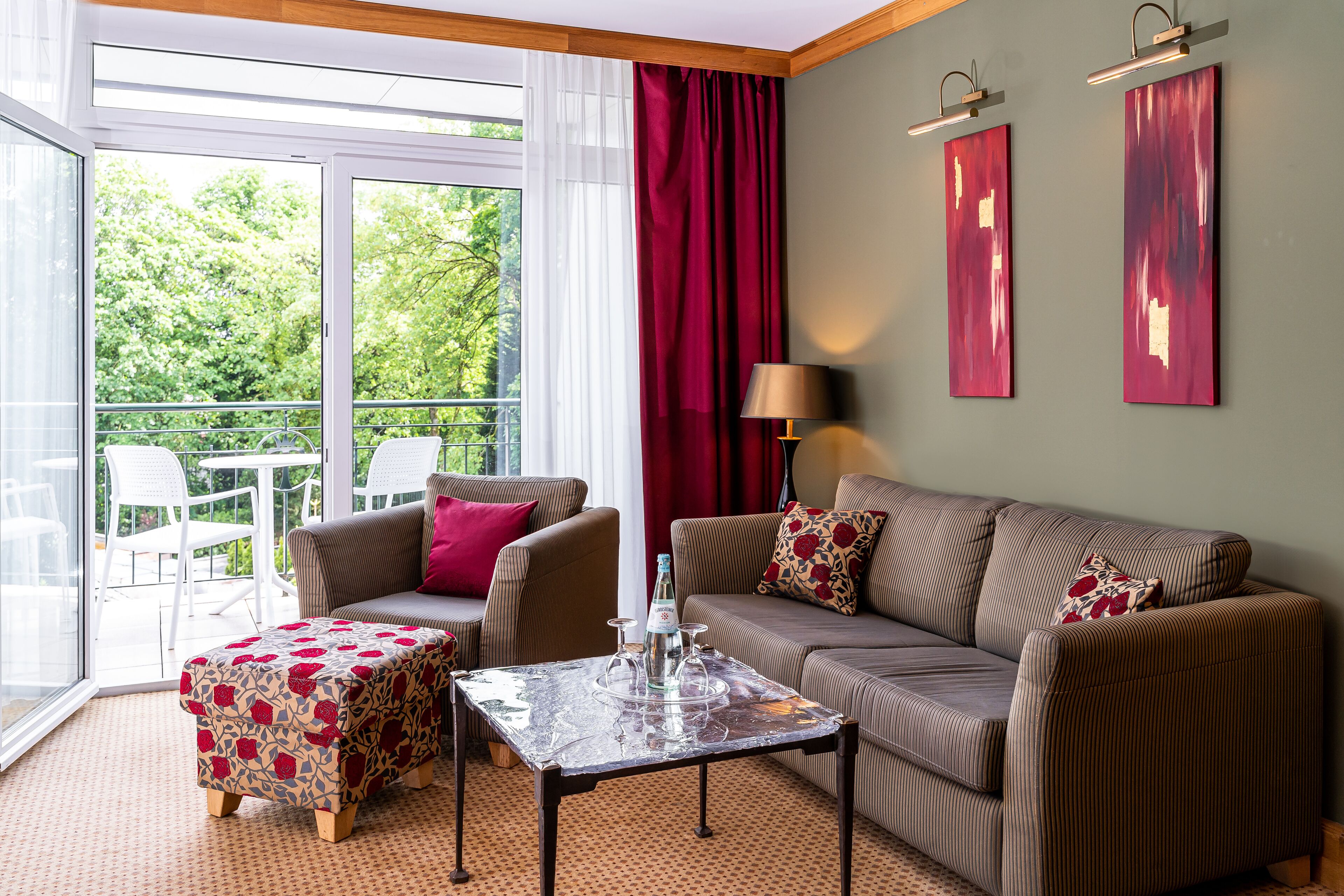 Ein modernes Wohnzimmer mit Sofa, Hocker und Tisch, ein Balkon mit Sitzgelegenheiten und Blick ins Grüne.