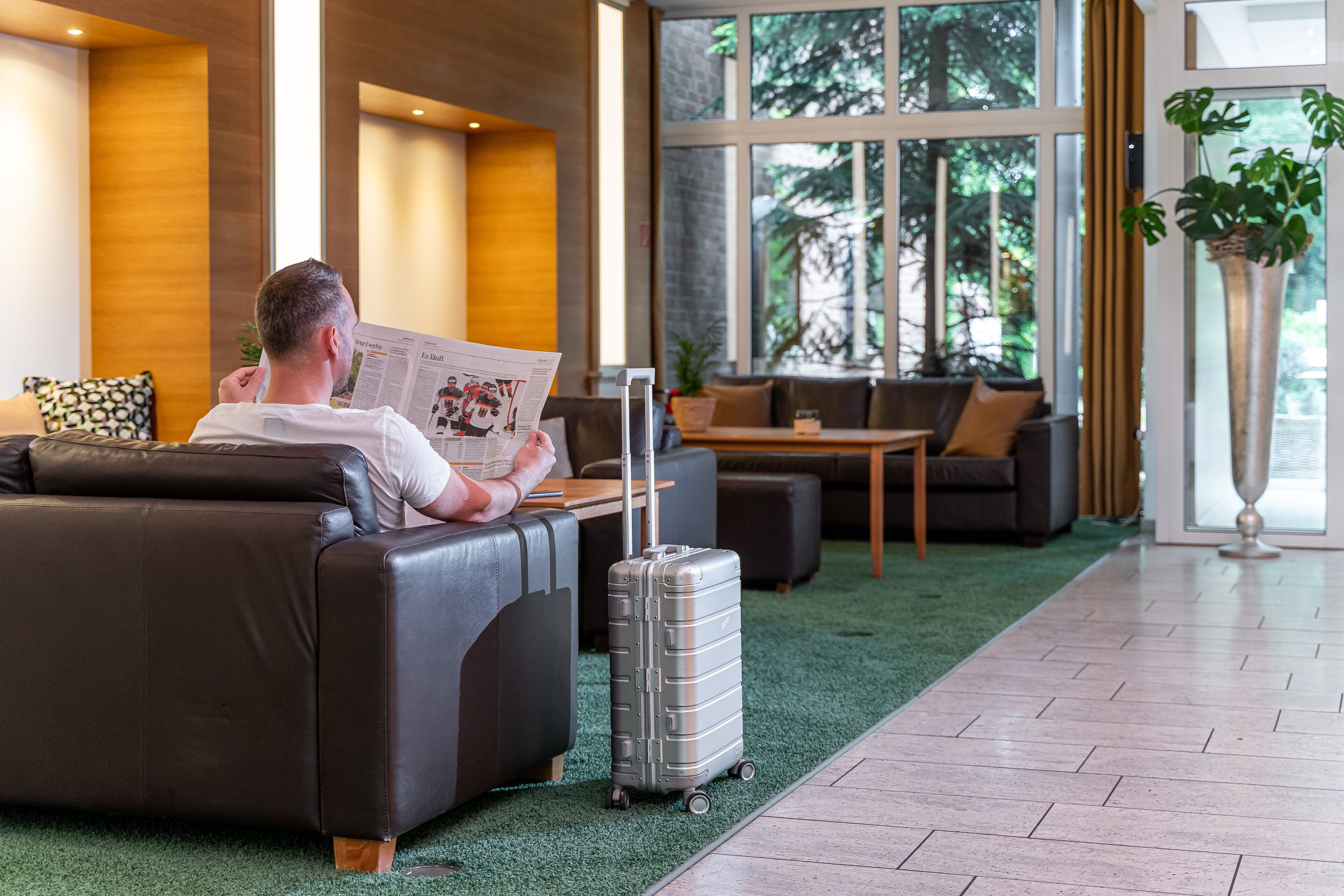 A man reading a magazine in a modern lounge area with leather sofas, a small table, and a potted plant.