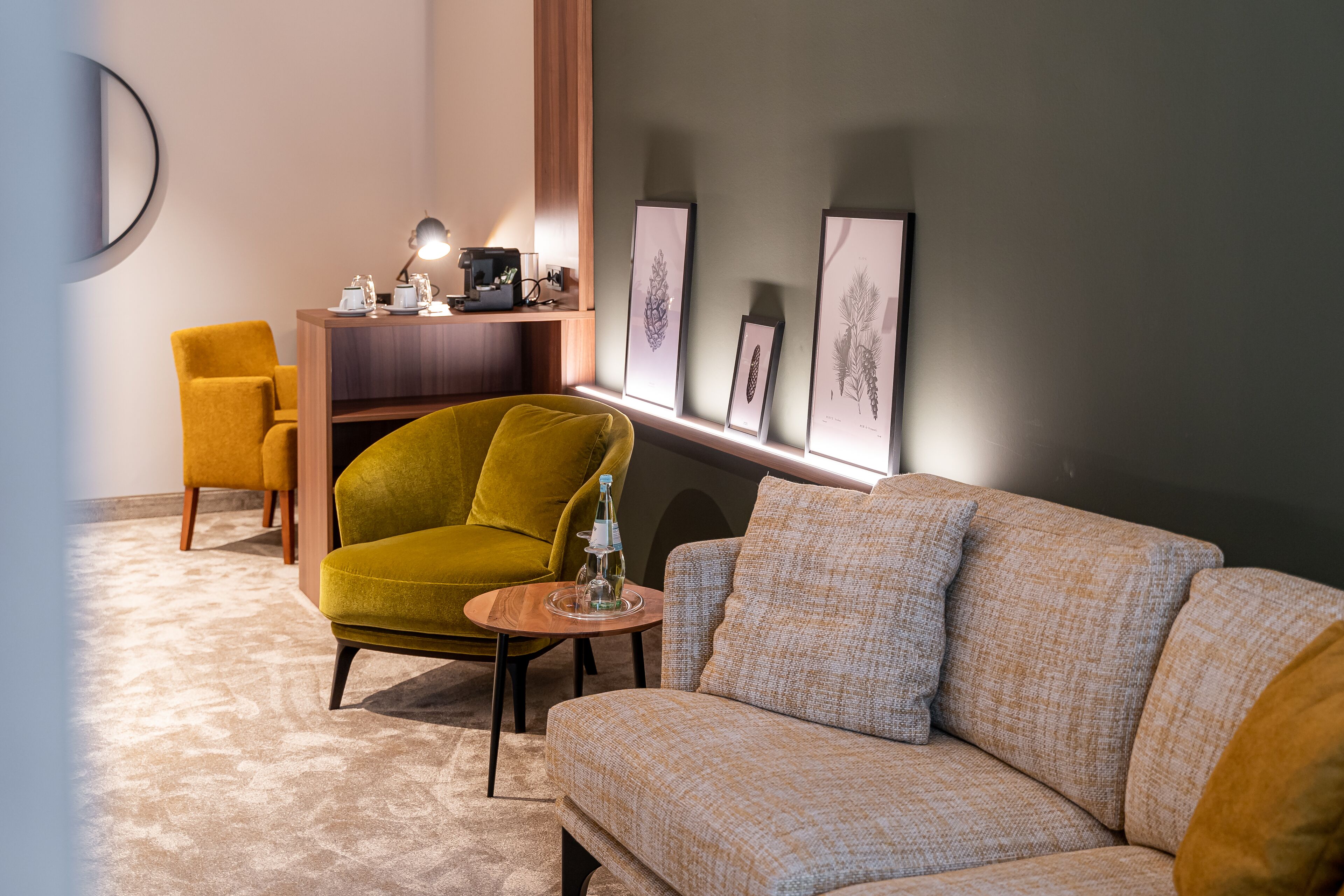 A modern living room with a sofa, a yellow armchair, a small wooden table with a bottle, and framed botanical prints on the wall.