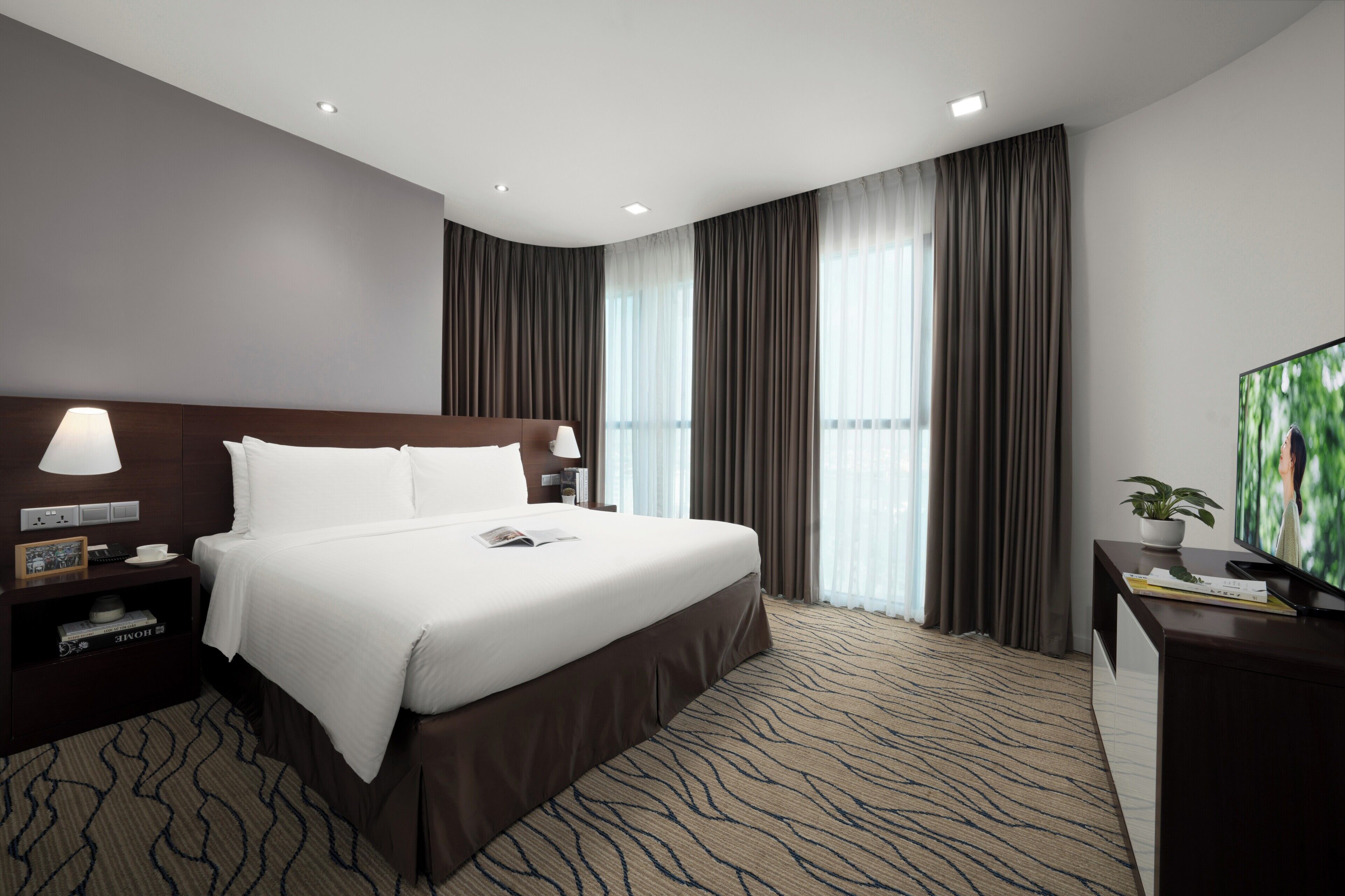 Et moderne hotelværelse med en stor seng, et fjernsyn, et skrivebord med en plante og udsigt til det frie gennem vinduerne.