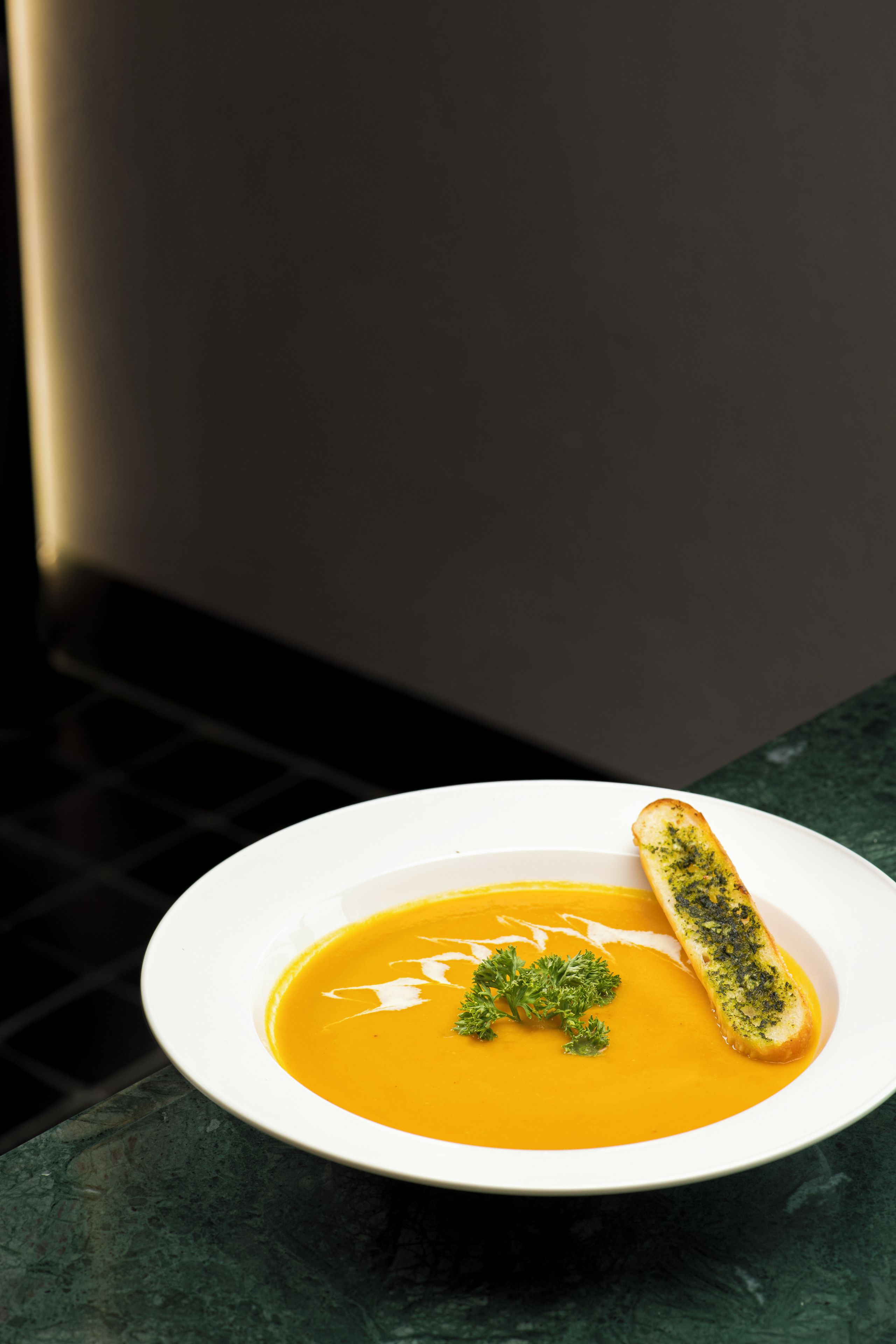 A white bowl of soup with a piece of bread on the side, placed on a dark surface.