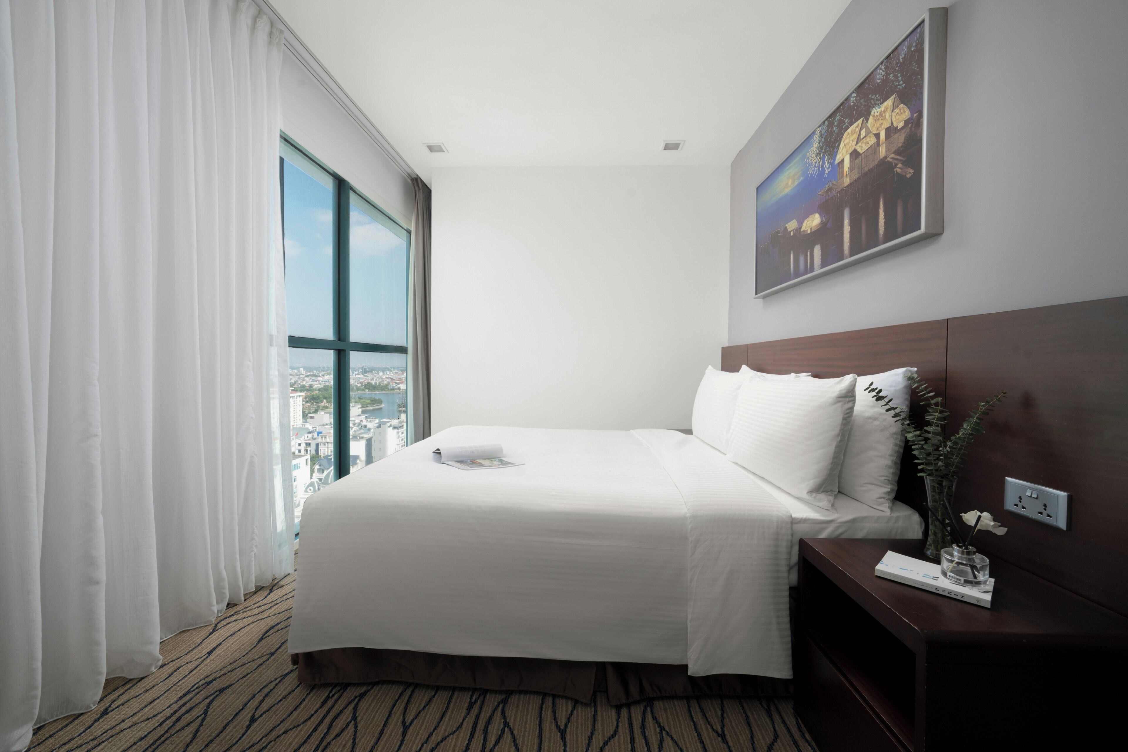 A hotel room with a large bed, a nightstand, a painting on the wall, and a view through the window.