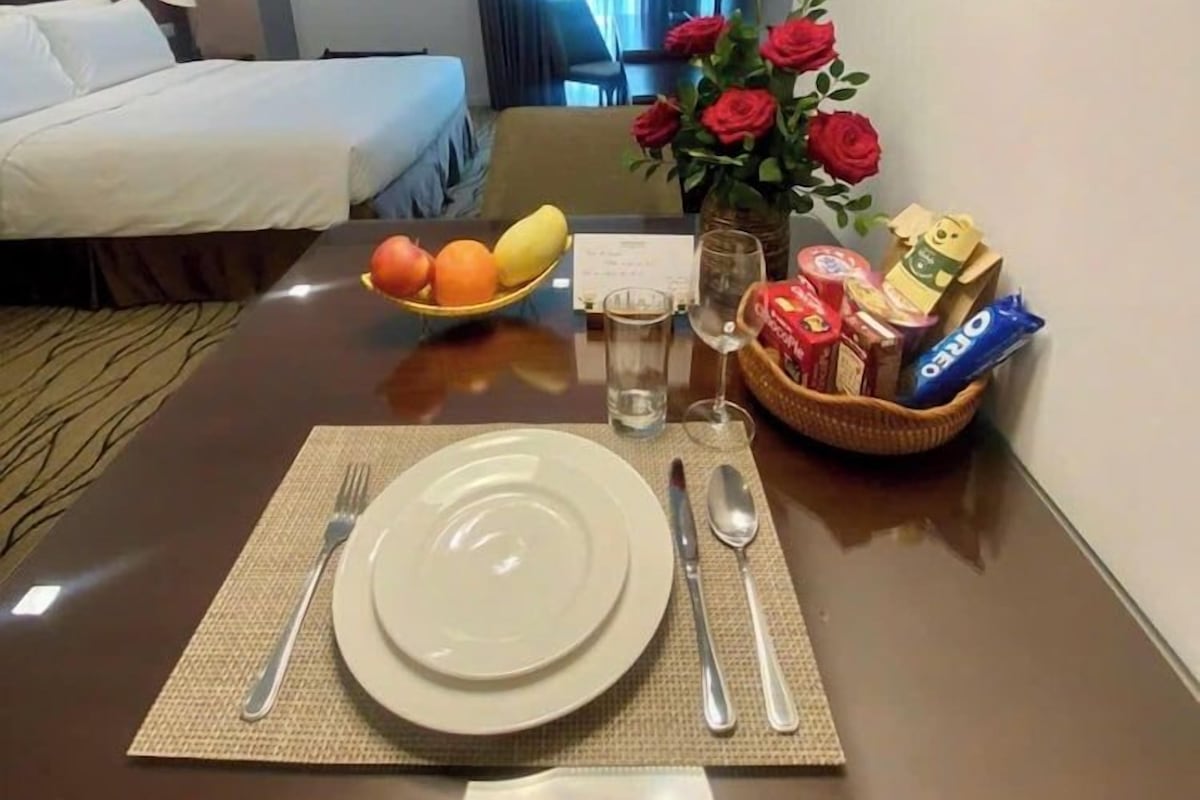 Une chambre d’hôtel avec un lit soigneusement fait, une table à manger dressée avec une assiette, des couverts et un panier de fruits.