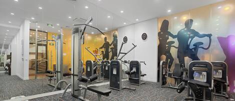 Una palestra con diverse macchine per esercizi, un tappeto a motivi e una parete con sagome di persone che si allenano.