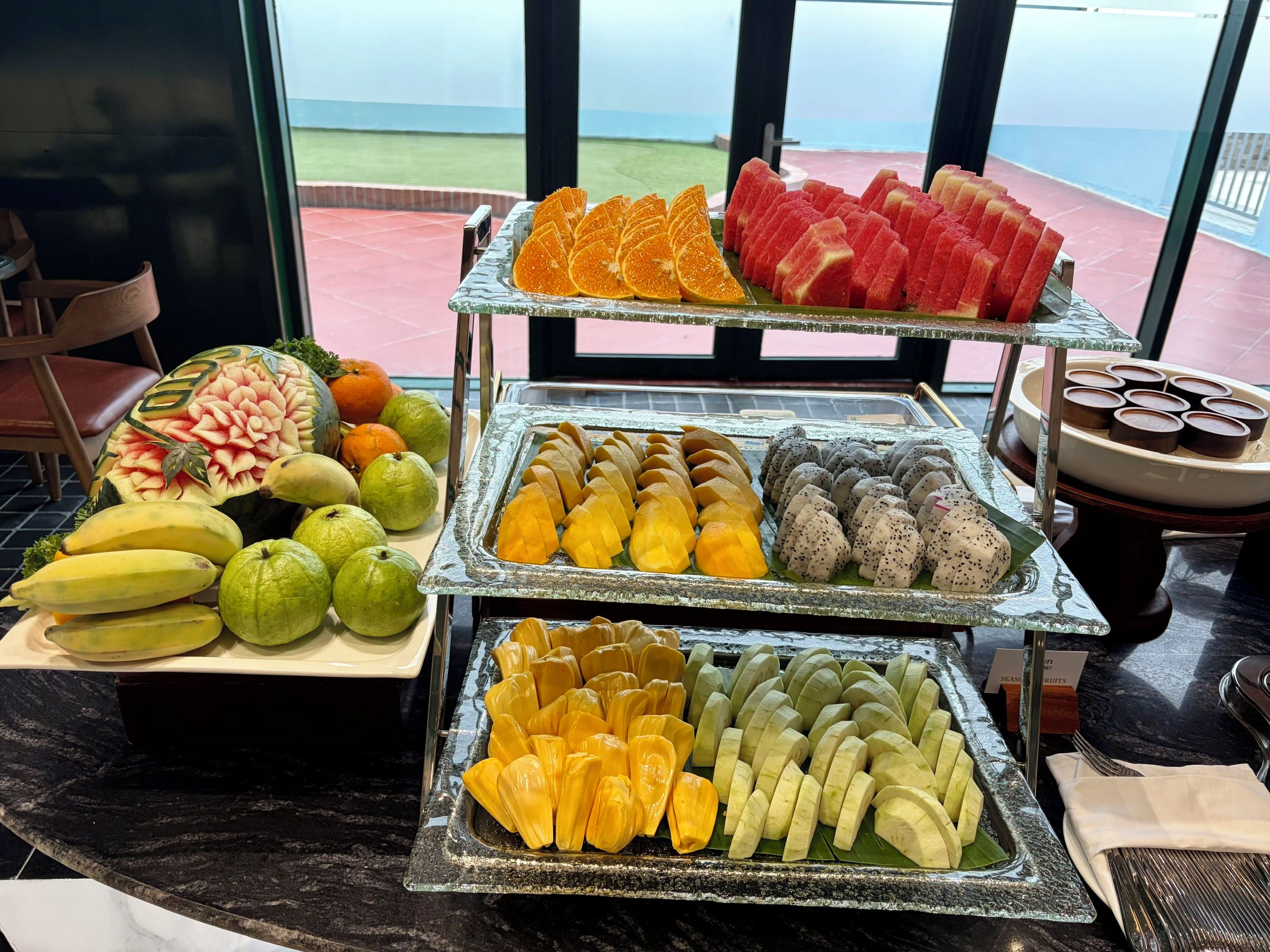 A fruit display with various types of fruits and a view of the sea through large windows.
