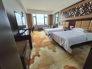 A hotel room with two beds, a desk, a TV, and a view of the city. - Manhattan International Hotel (Huaibei)
