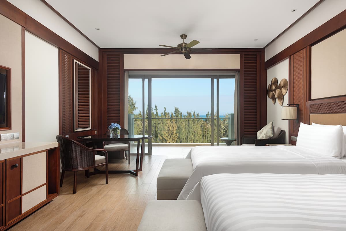 A hotel room with two beds, a desk, a chair, and a balcony with a view.