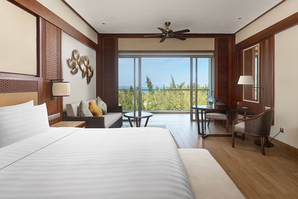 A hotel room with a large bed, a sofa, a small table, and a chair. There is a balcony with a view of the ocean.